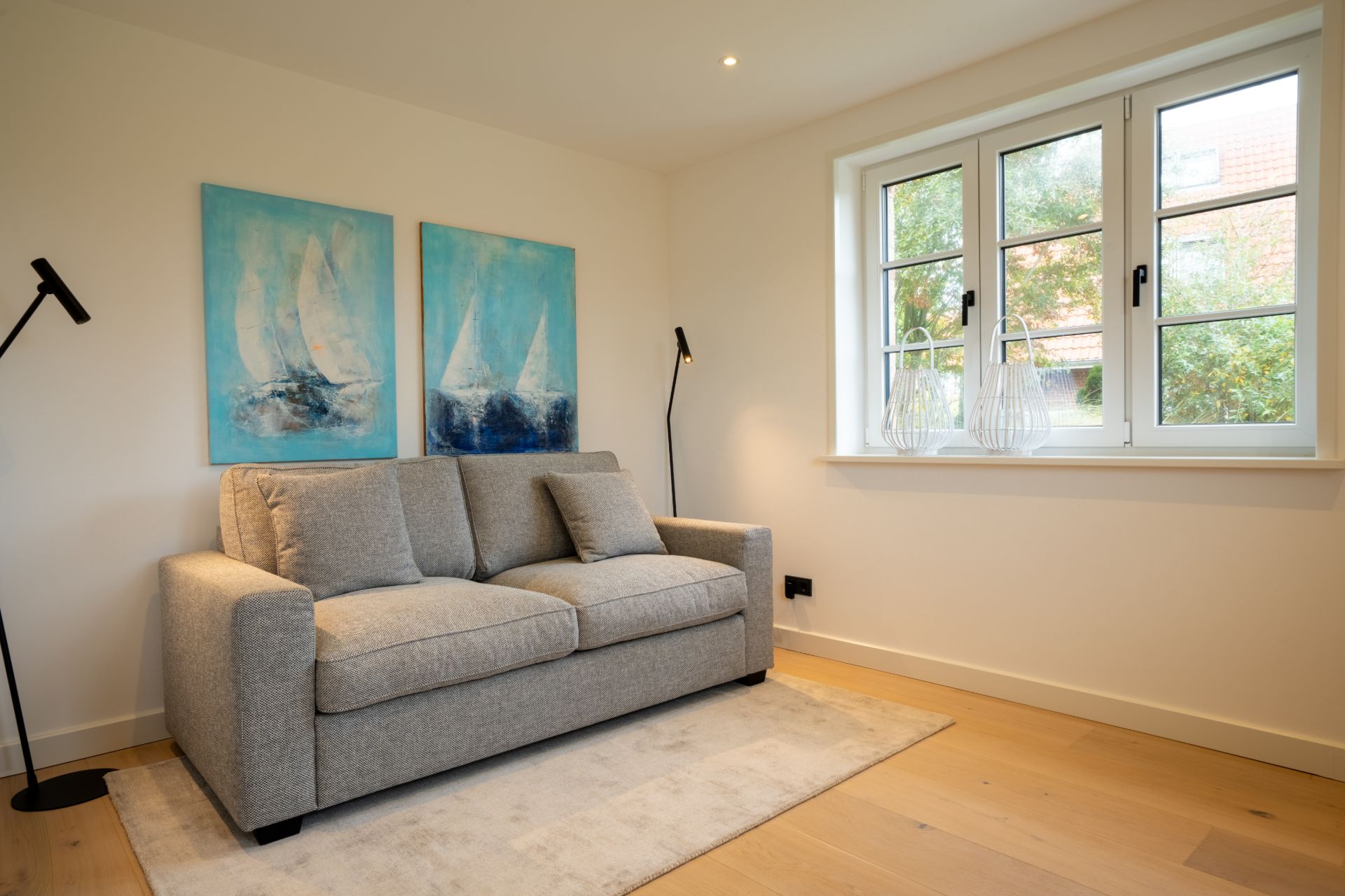 Helles Wohnzimmer mit grauem Sofa, großem Fenster und zwei Bildern von Segelbooten an der Wand.