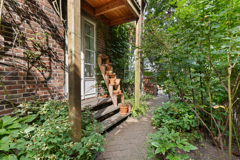 Kleiner Hauseingang mit Holztreppe, Blumentöpfen und umgeben von viel Grün.