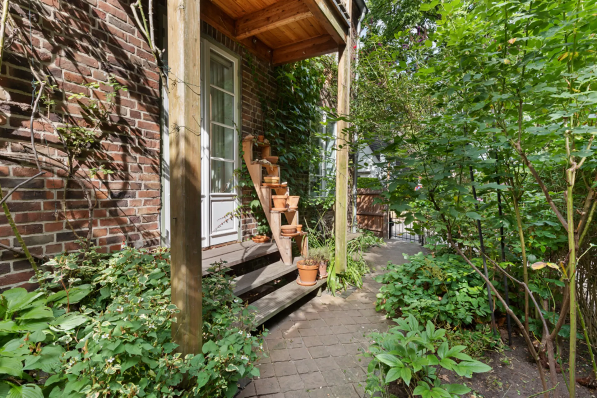 Kleiner Hauseingang mit Holztreppe, Blumentöpfen und umgeben von viel Grün.