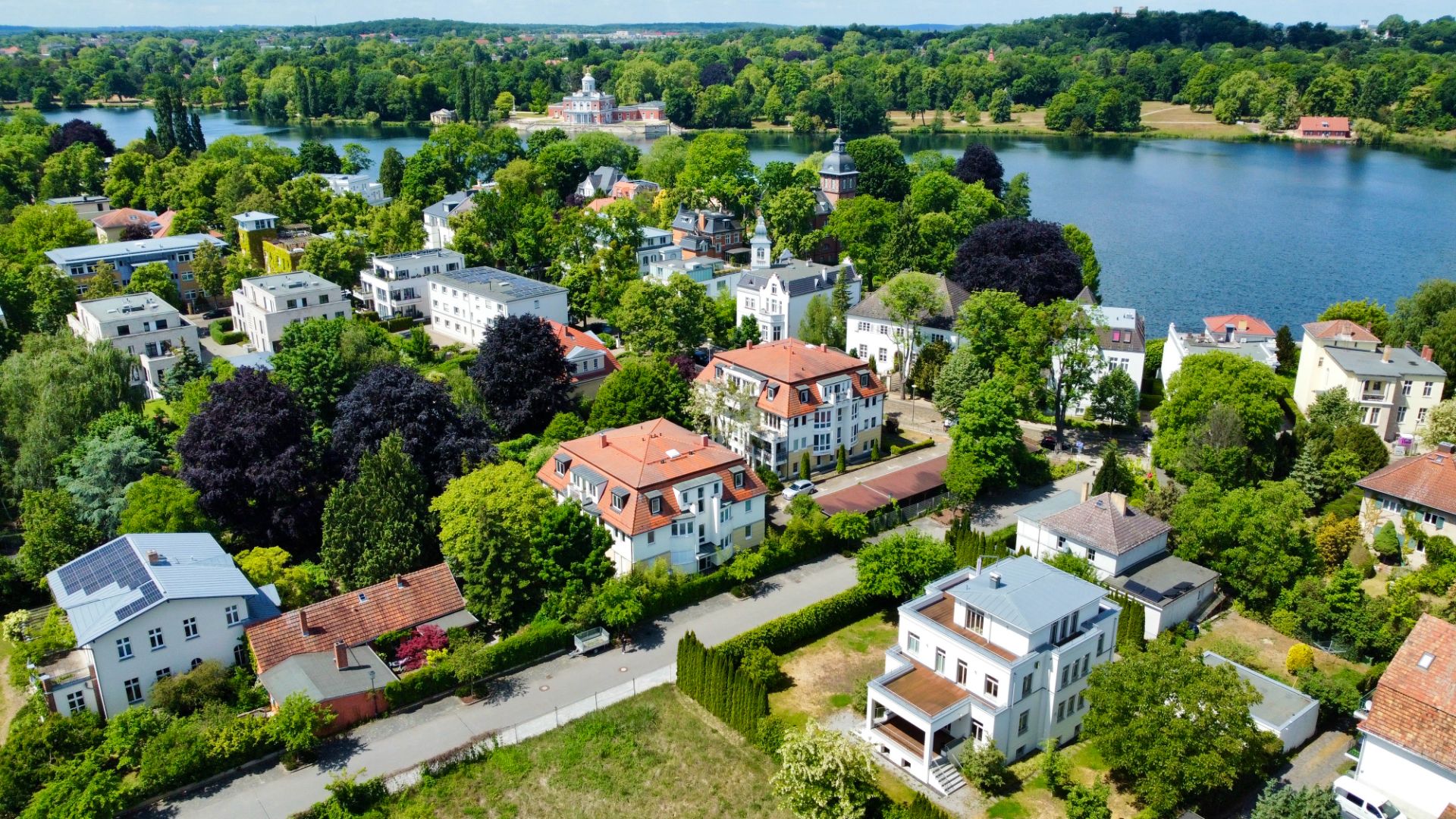 Luftaufnahme von Wohnhäusern mit roten und weißen Dächern, Bäumen und einem See im Hintergrund.