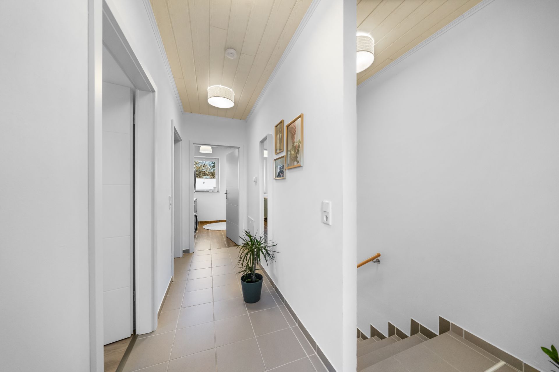 Heller Flur mit Fliesenboden, Zimmern, Treppe, Pflanzen und Bildern an der Wand.