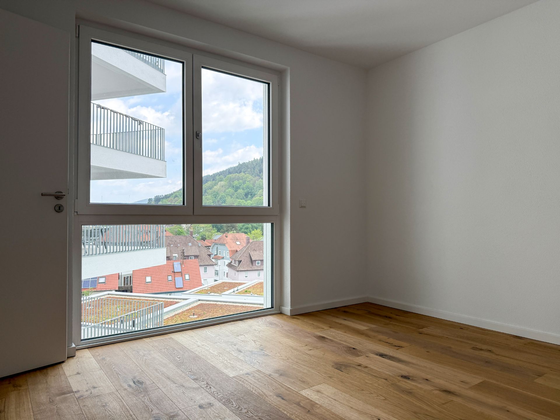 Leeres Zimmer mit Holzboden und großem Fenster mit Blick auf Balkone und Häuser.