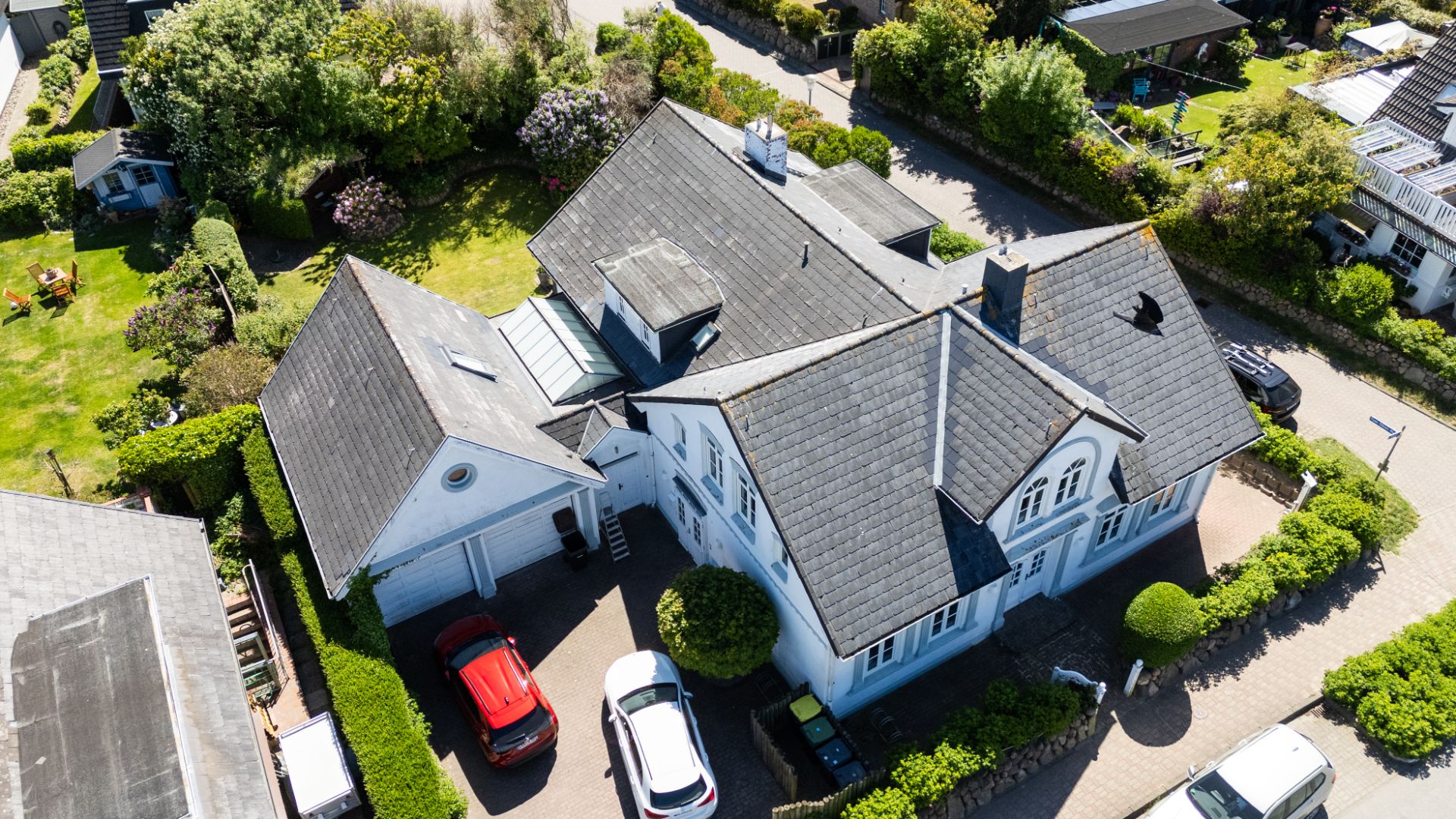 Luftaufnahme eines großen weißen Wohnhauses mit mehreren Autos in der Einfahrt und Garten rundherum.