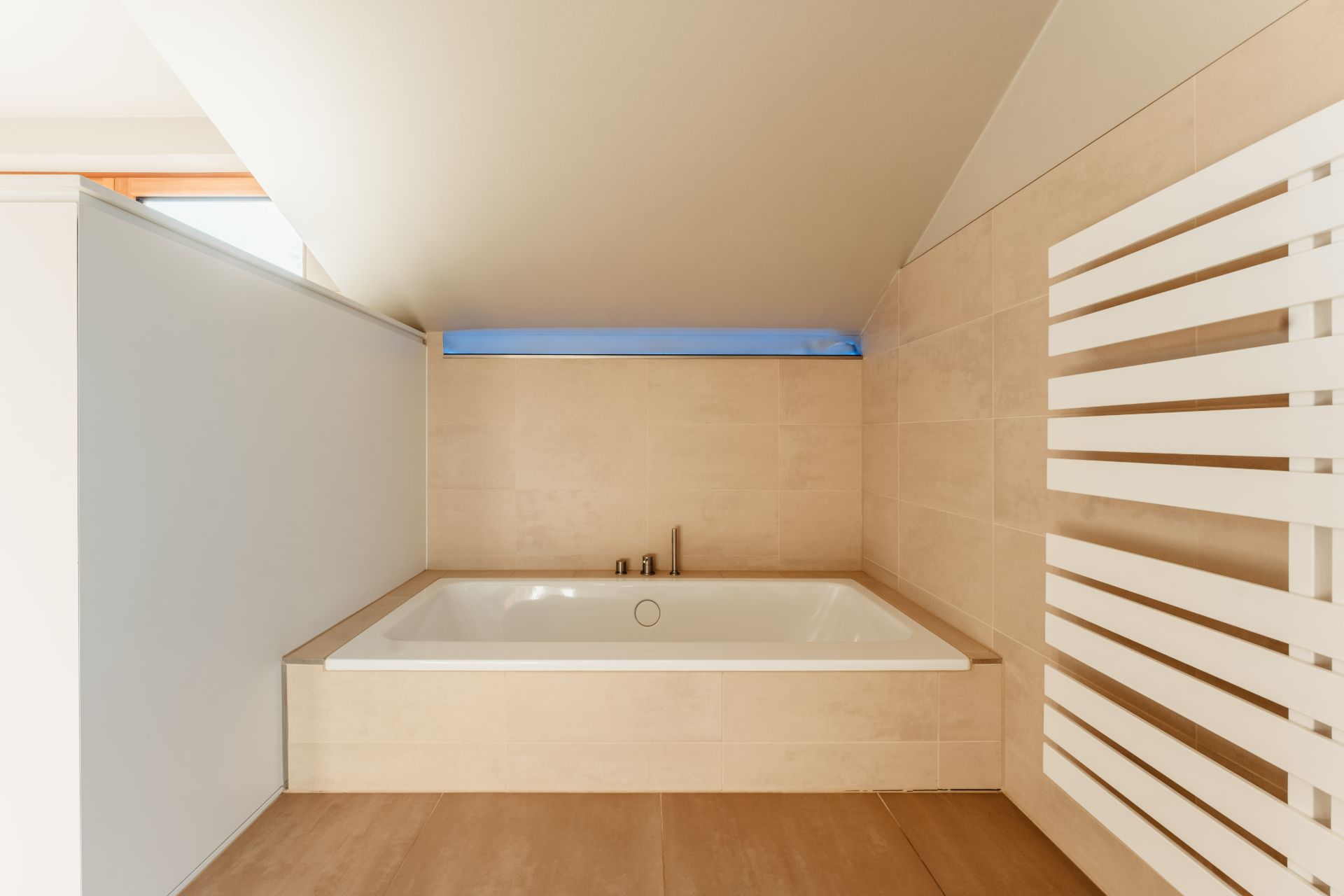 Bad mit Badewanne unter einer Dachschräge, beigen Fliesen und modernem Heizkörper.