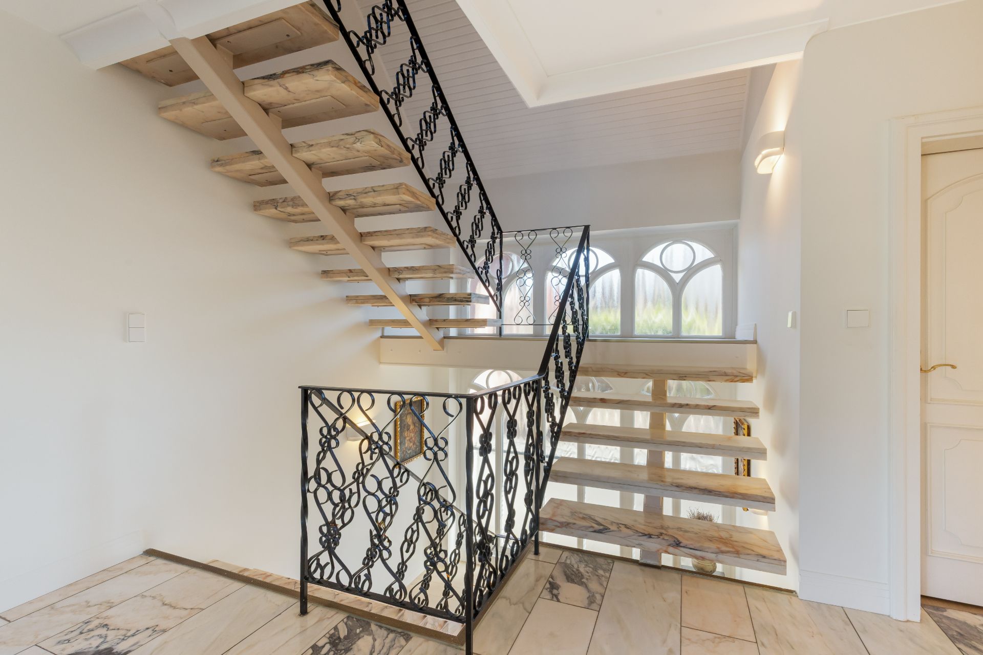 Moderne Holztreppe mit verziertem schwarzen Geländer in einem hellen Flur mit Marmorboden.