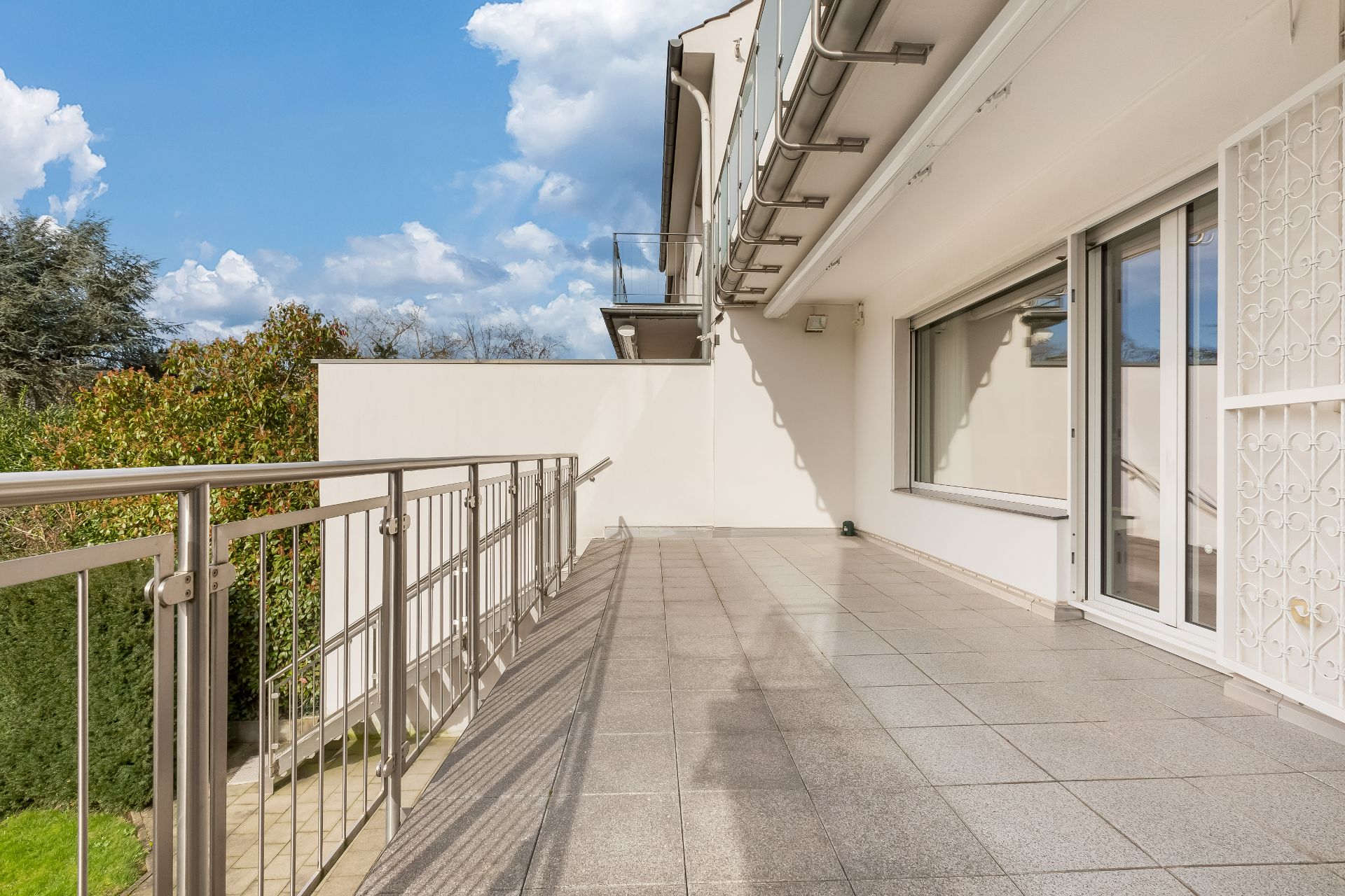 Große Terrasse mit Fliesenboden, Metallgeländer und Zugang zu einem Garten.