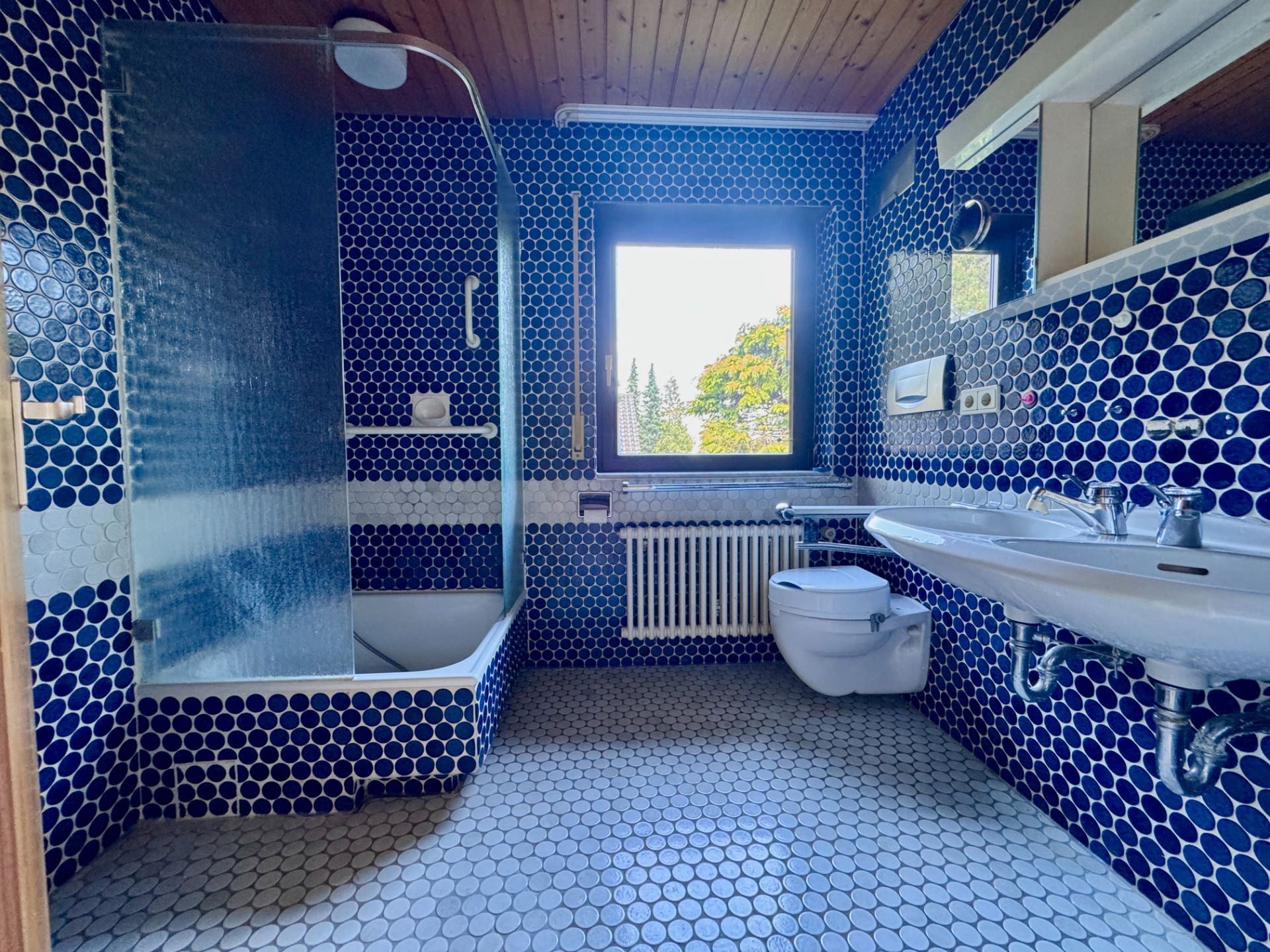 Badezimmer mit blauen Mosaikfliesen, Badewanne, Waschbecken, WC und großem Fenster.