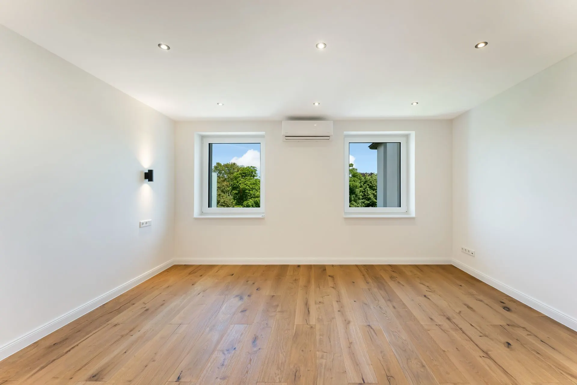Leerer Raum mit Holzboden, zwei Fenstern und einer Klimaanlage an der Wand.