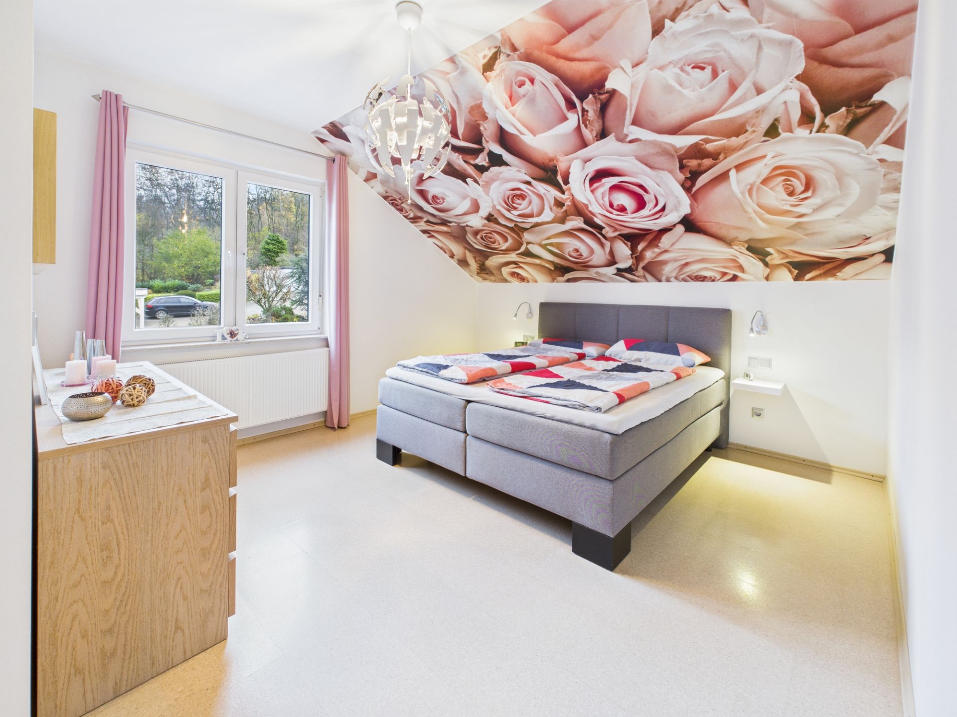 Schlafzimmer mit grauem Bett, Kommode und großem Rosenmotiv an der Dachschräge.