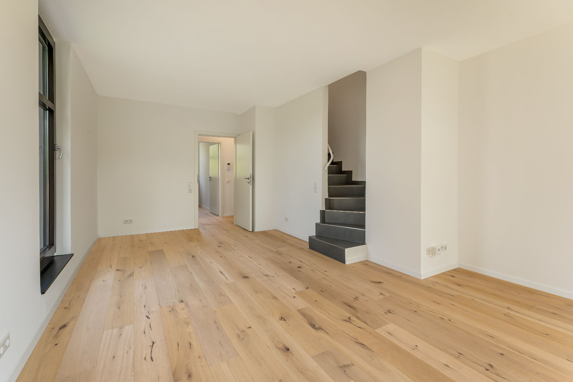 Helles Wohnzimmer mit Holzboden, großem Fenster und einer Treppe nach oben.