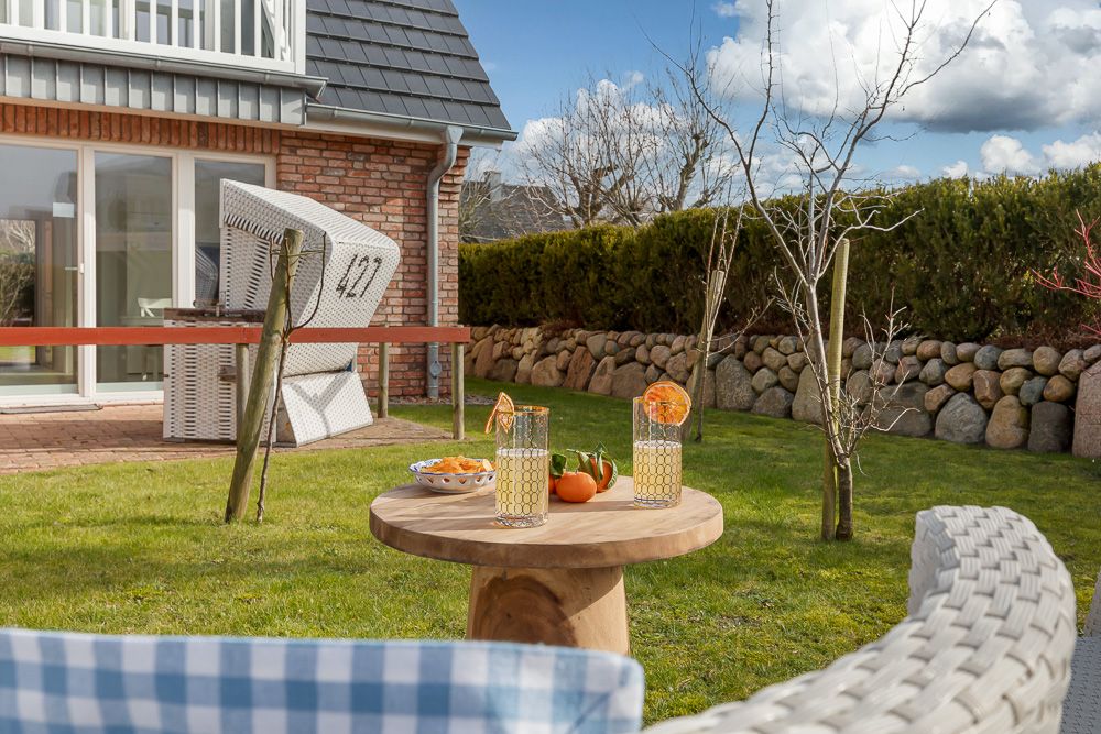 Garten mit Holztisch, zwei Getränken, Obstschale und Strandkorb vor Haus mit Steinmauer.