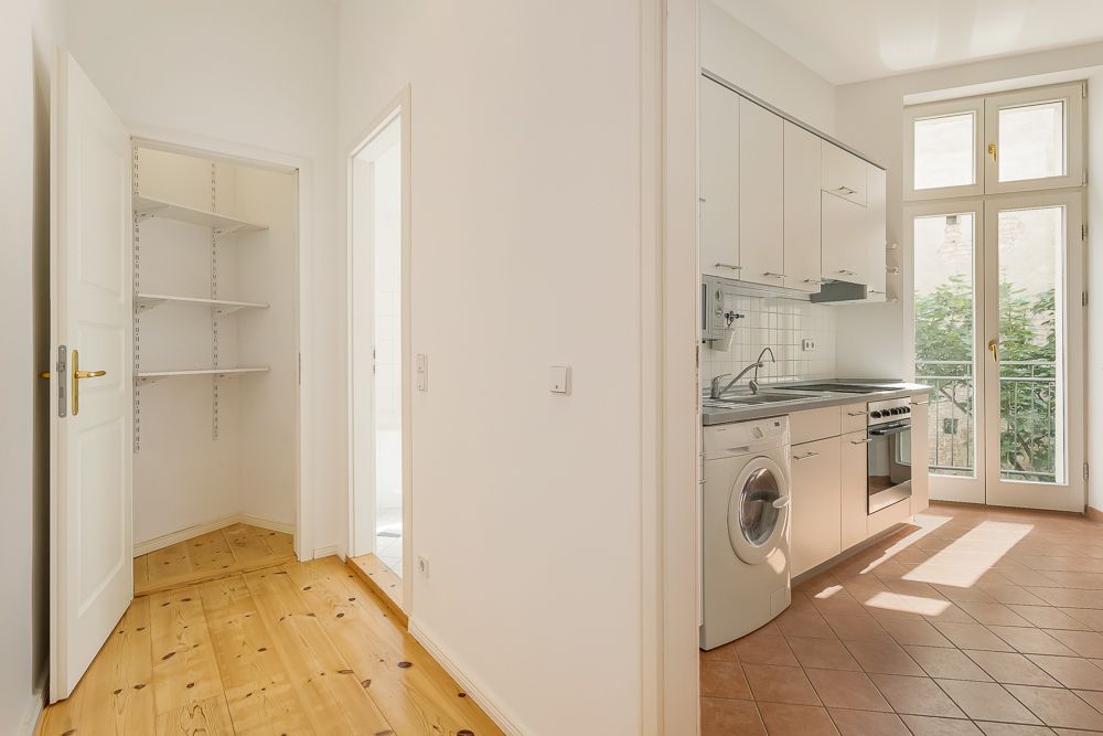 Helle Küche mit Einbauschränken, Herd, Waschmaschine und Zugang zum Balkon. Links ein Abstellraum mit Regalen.