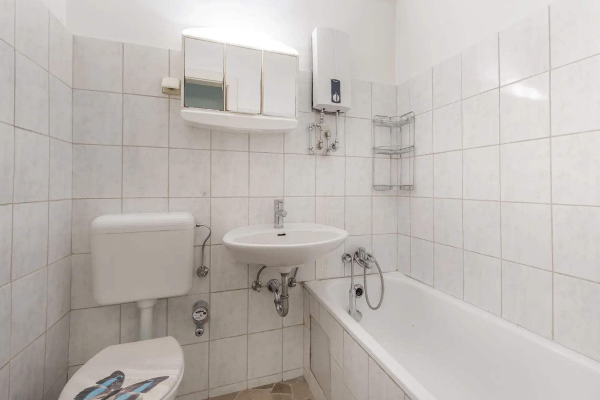 Weiß gefliestes Badezimmer mit Toilette, Waschbecken, Spiegel, Badewanne und Wandregal.
