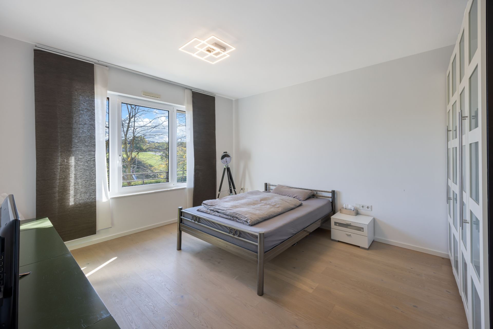 Helles Schlafzimmer mit großem Fenster, Bett, Nachttisch und Kleiderschrank.