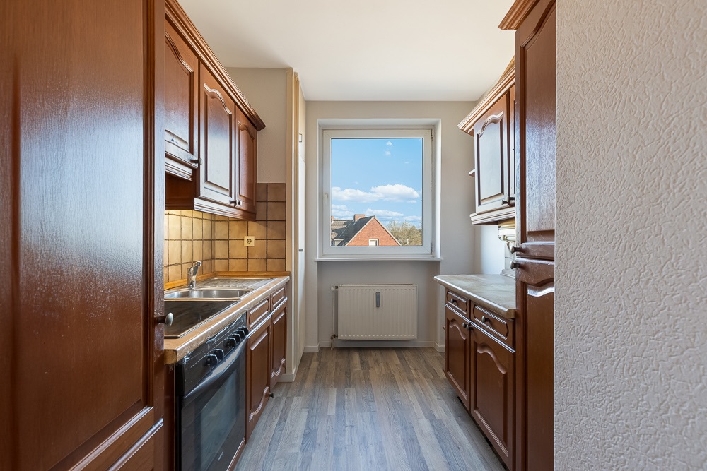 Schmale Küche mit Holzschränken, braunen Fliesen und Fenster mit Blick ins Freie.