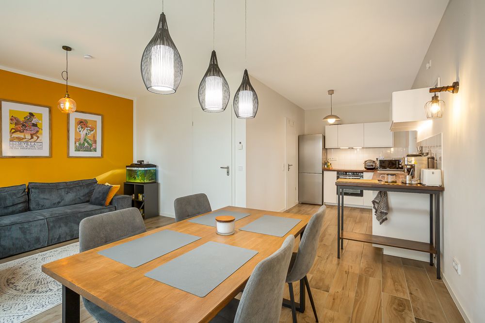 Wohnzimmer mit Esstisch, grauem Sofa, gelber Wand, Aquarium und offener Küche, heller Holzboden.