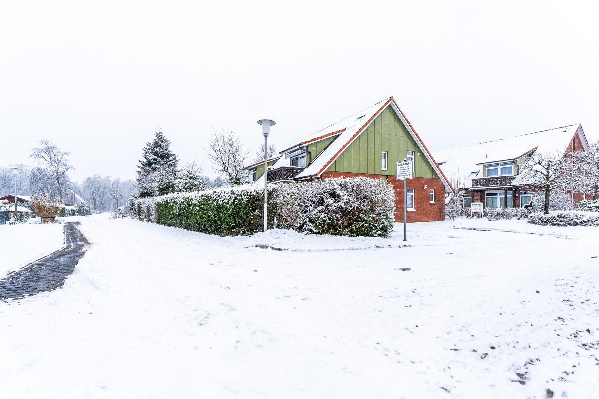 Wohnhäuser und Gehweg mit Schnee bedeckt, Hecken und Bäume im Winter.