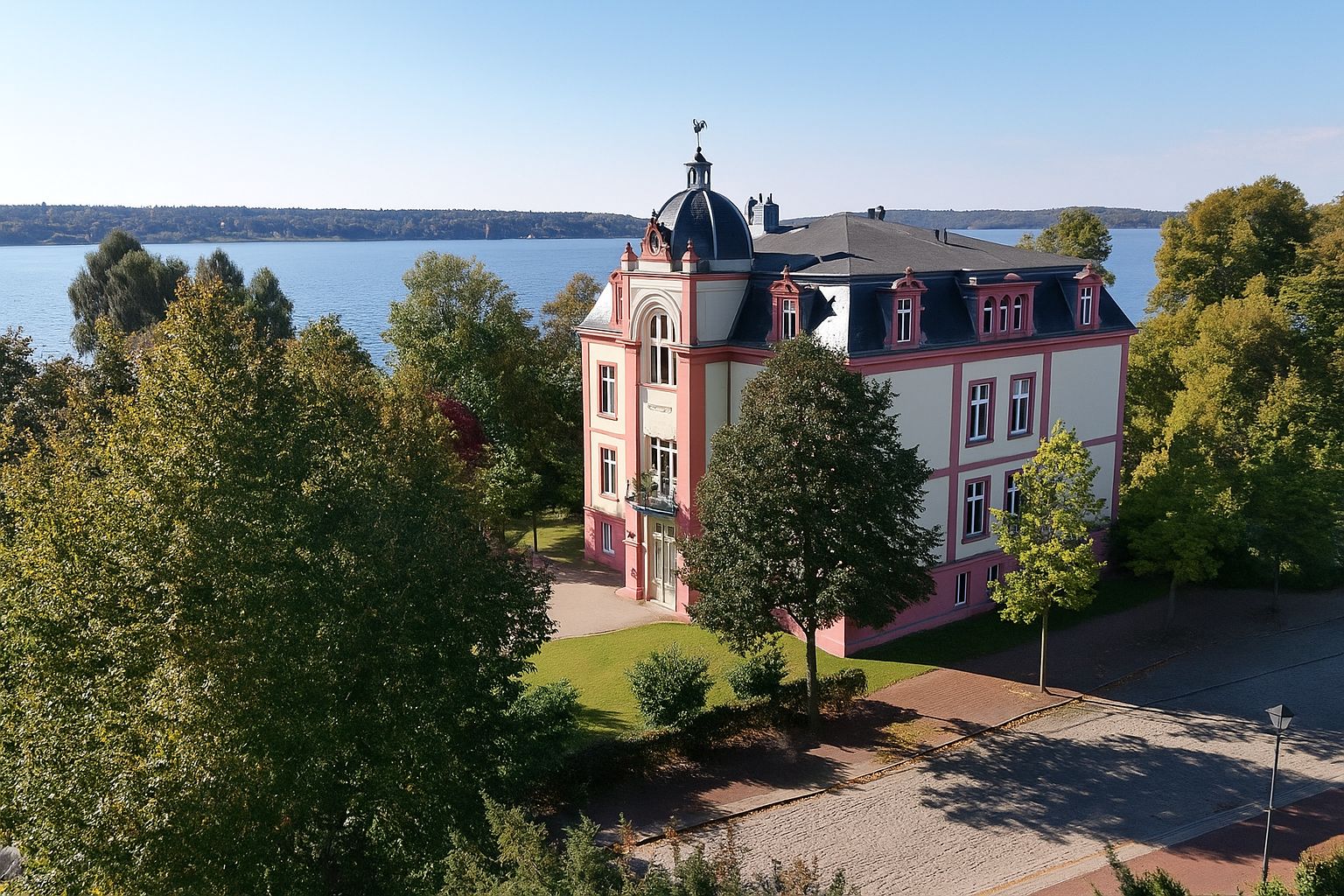 Großes, mehrstöckiges Wohnhaus mit Kuppeldach am See, umgeben von Bäumen und Straße.
