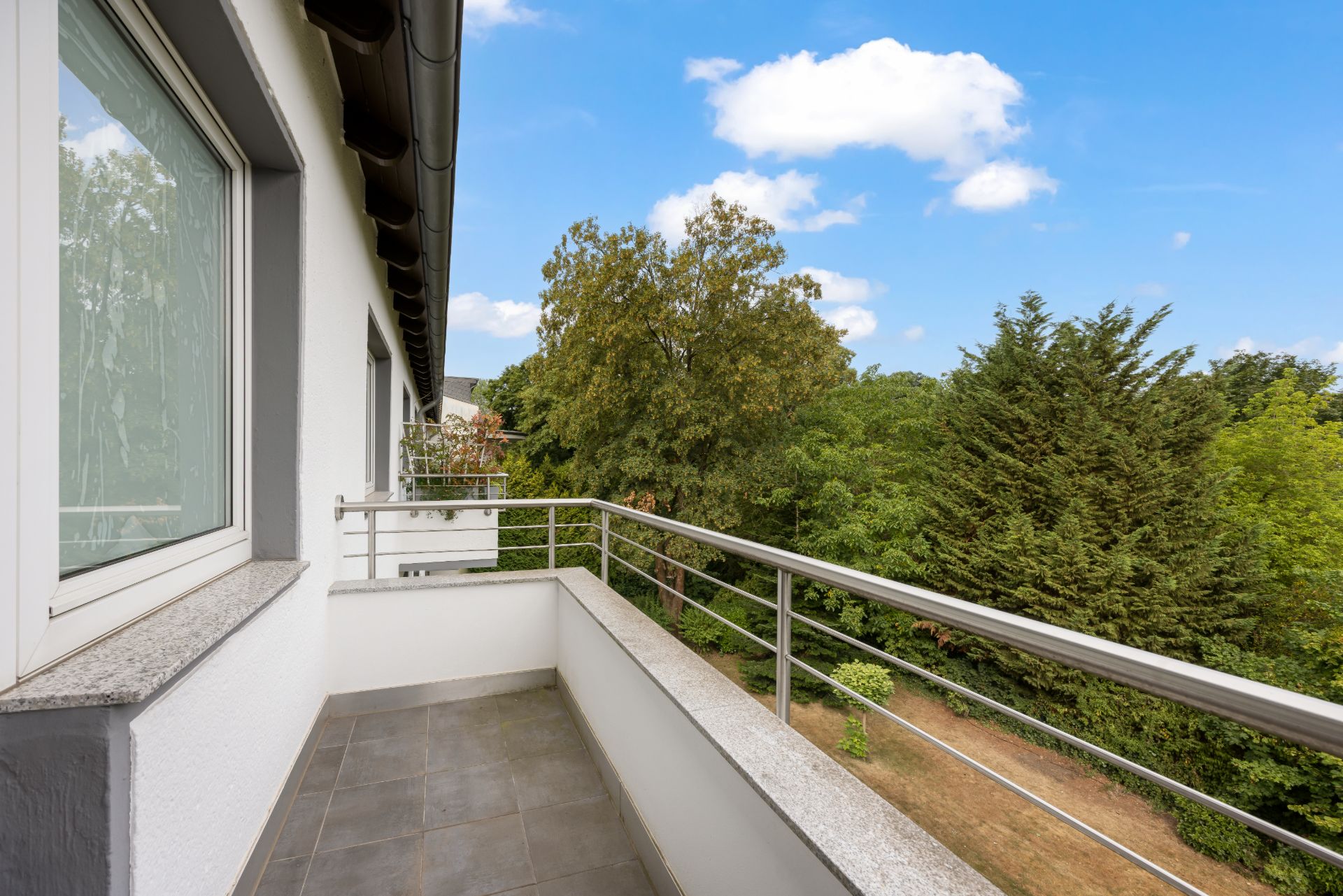 Balkon mit Metallgeländer und Blick auf grüne Bäume und blauen Himmel.