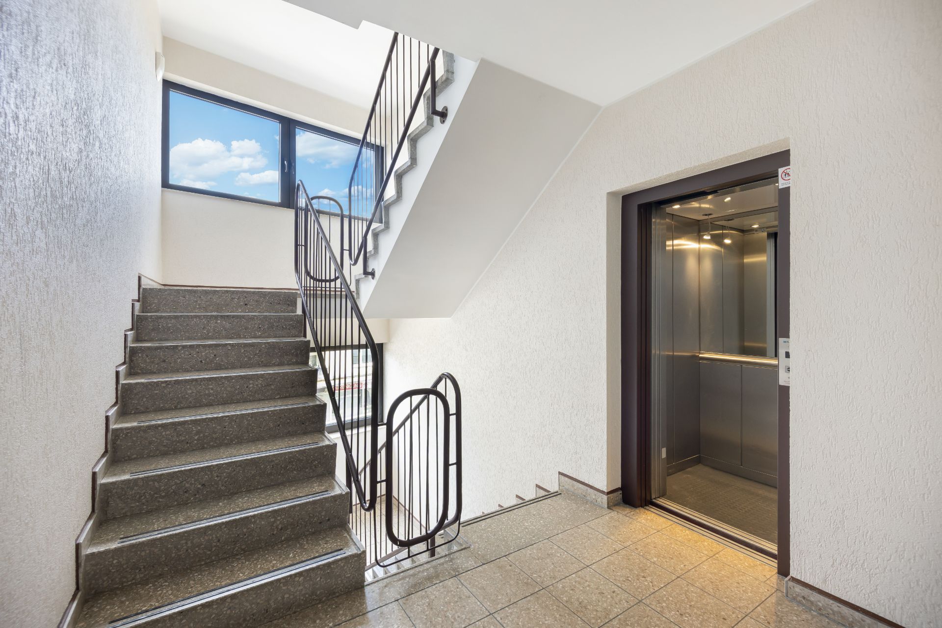 Treppenhaus mit Aufzug und großem Fenster, Fliesenboden und Metallgeländer.