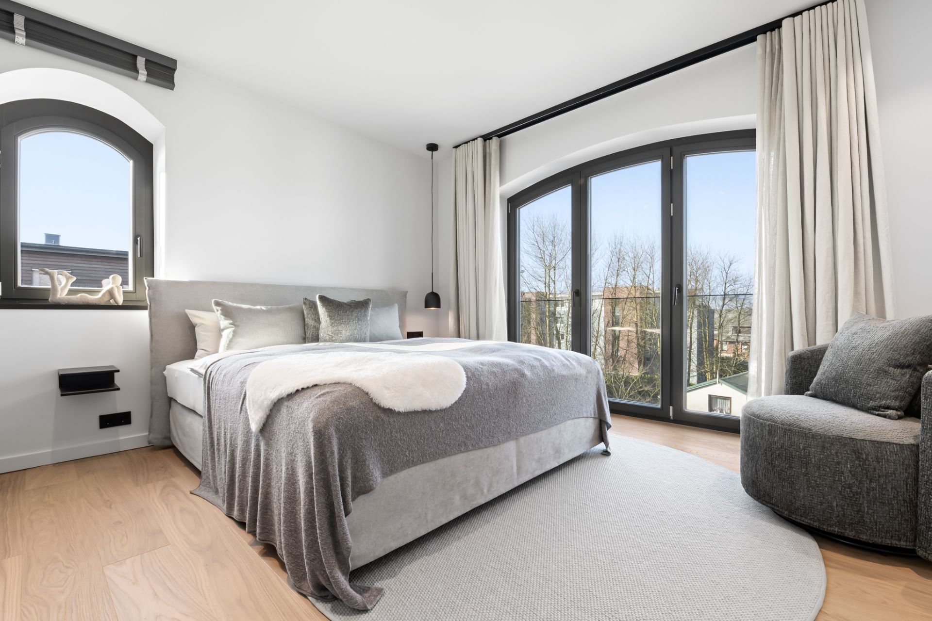 Helles Schlafzimmer mit großem Bett, Sessel und großen Fenstern mit Vorhängen.