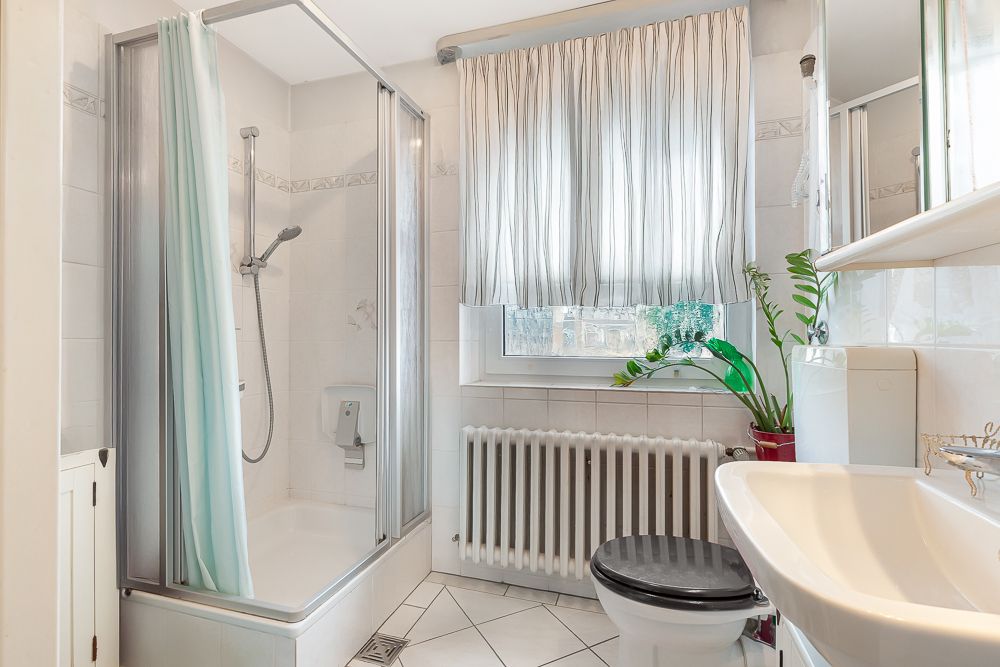 Helles Badezimmer mit Dusche, WC, Waschbecken, Fenster mit Vorhang und einer grünen Pflanze.