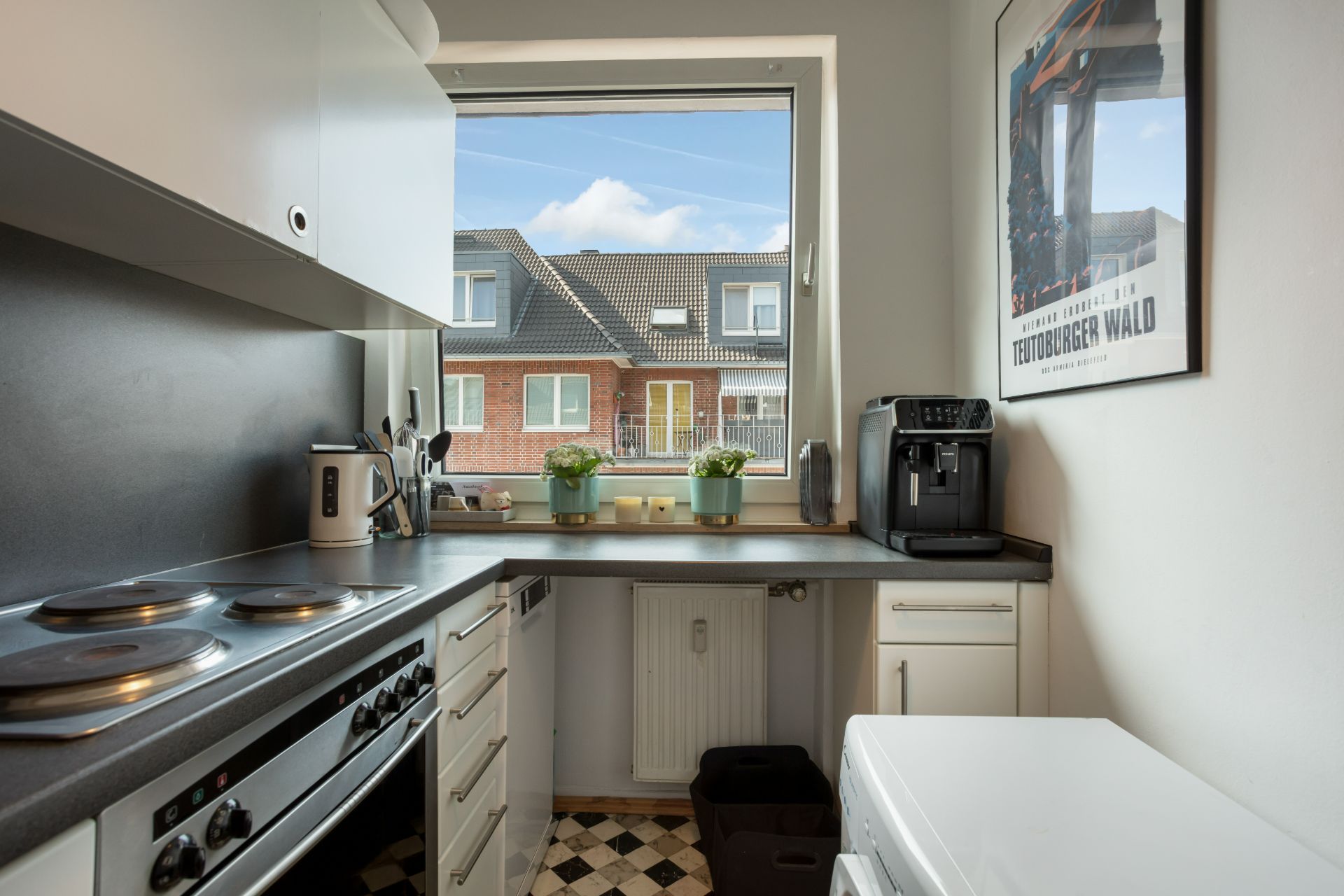 Kleine, helle Küche mit Herd, Kaffeemaschine, Fenster und Blick auf Nachbarhäuser.