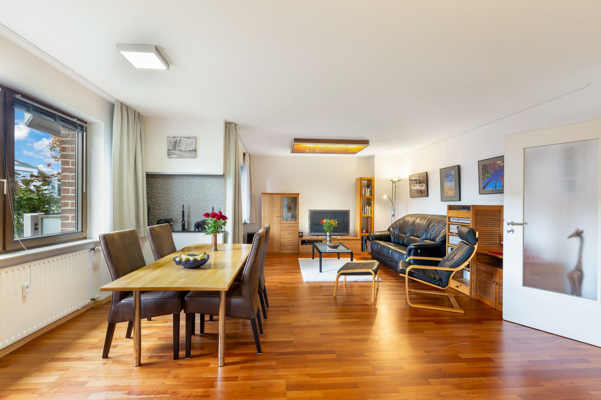 Helles Wohnzimmer mit Holzboden, Esstisch, Stühlen, Sofa und großen Fenstern.