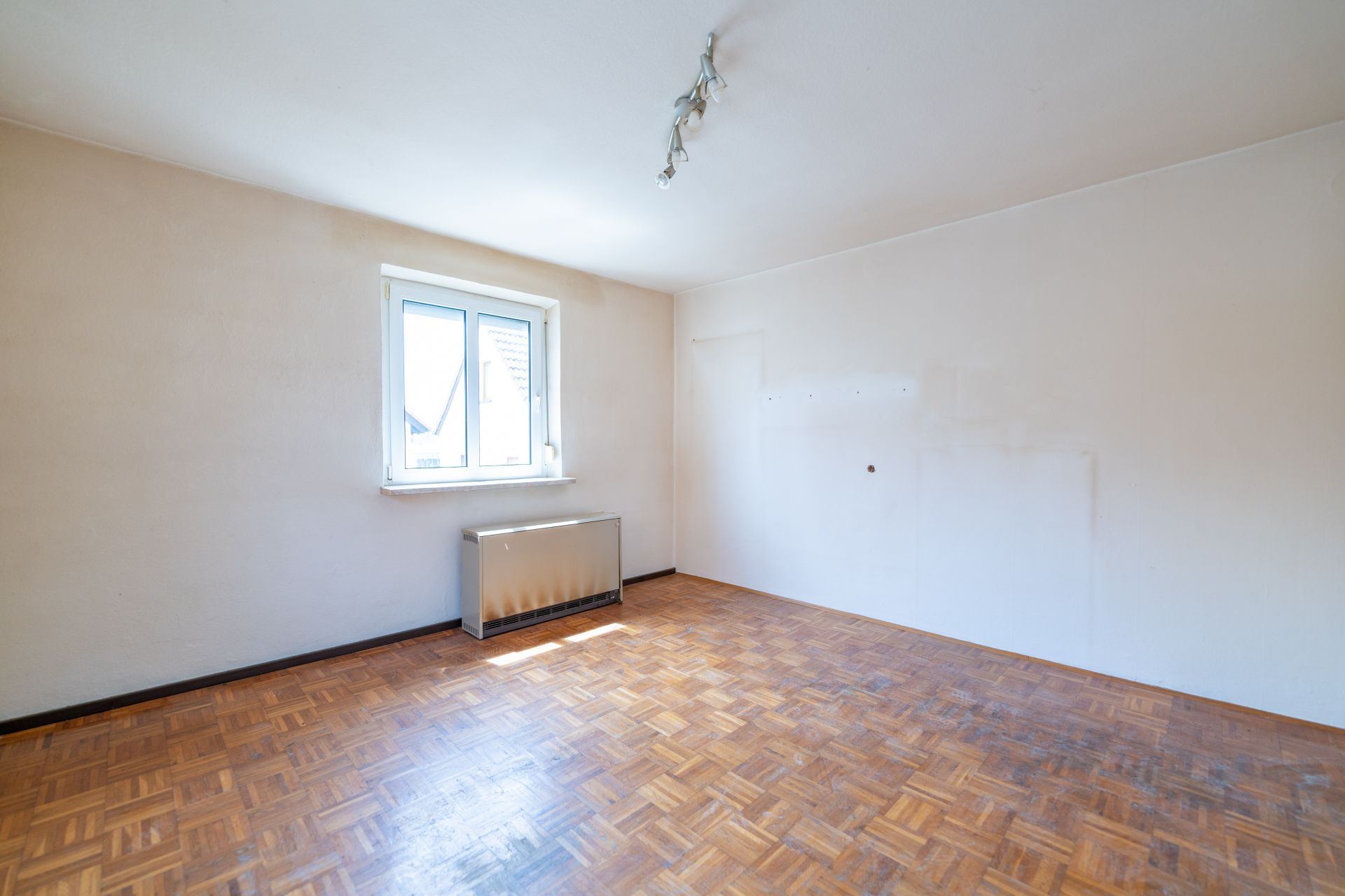 Leeres Zimmer mit Parkettboden, weißer Wand, Fenster und Heizkörper unter dem Fenster.
