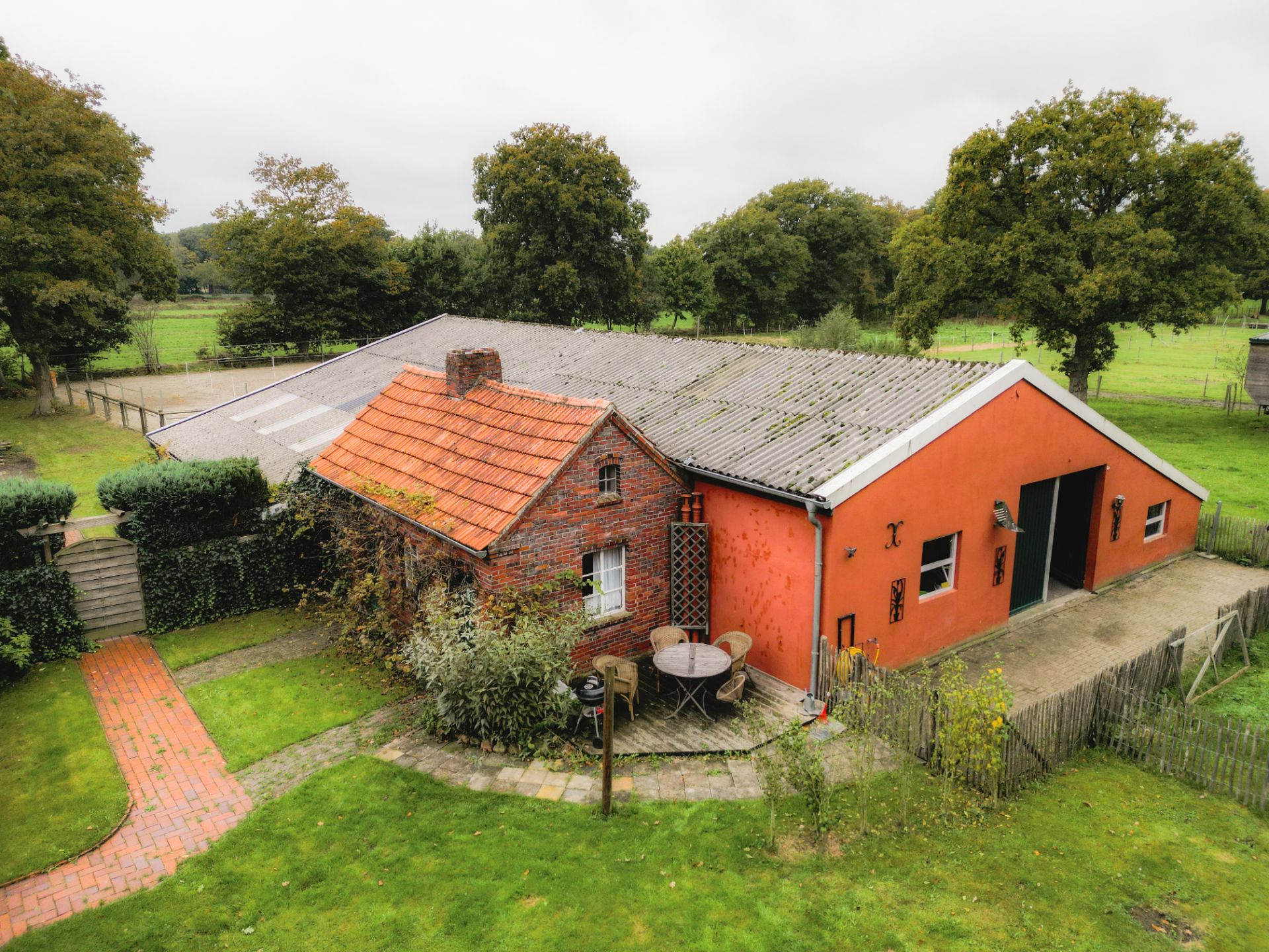 Backsteinhaus mit rotem Anbau, Terrasse mit Sitzgruppe und grünem Garten, umgeben von Bäumen.