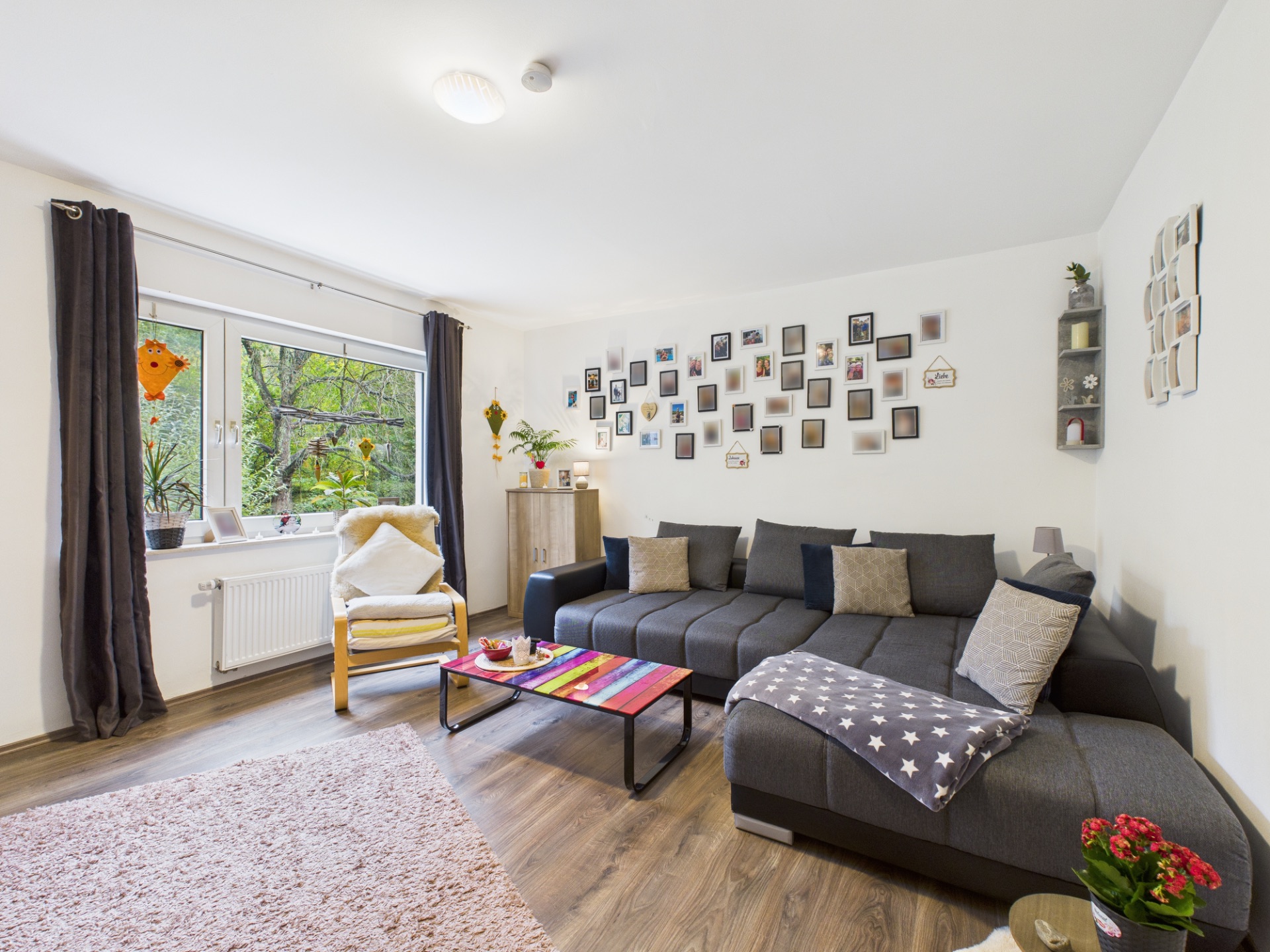 Modernes Wohnzimmer mit grauem Sofa, buntem Teppich, Sessel am Fenster und vielen Bildern an der Wand.