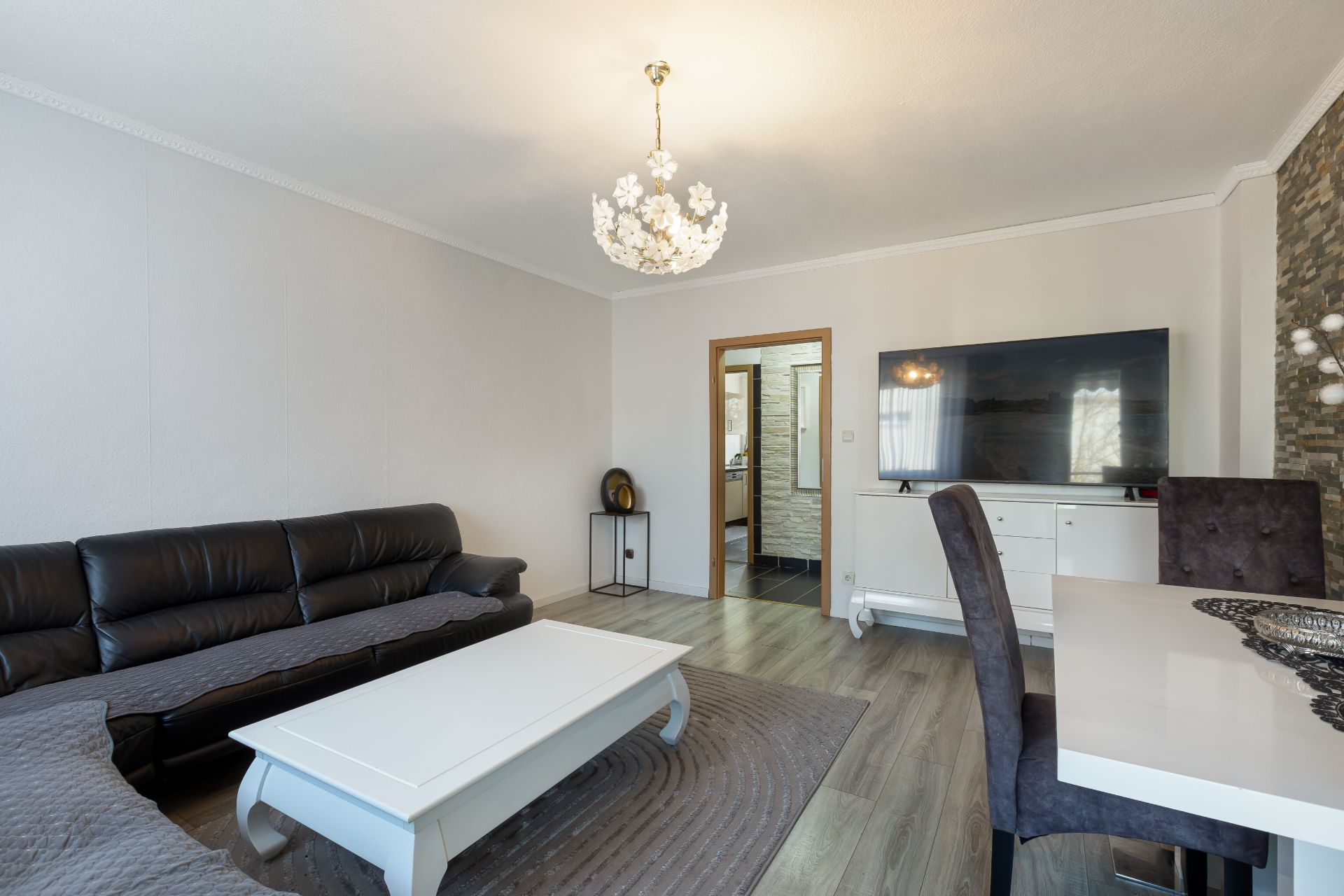 Wohnzimmer mit schwarzem Sofa, weißem Couchtisch, Esstisch, Fernseher und heller Lampe.