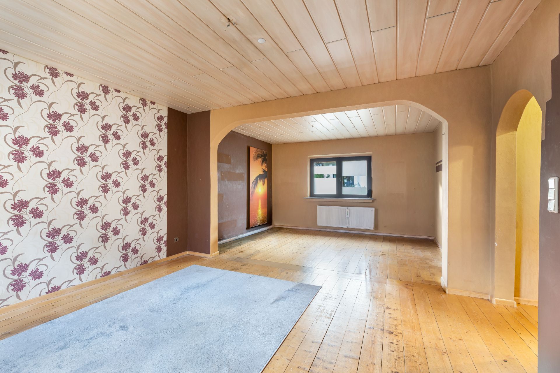 Helles Wohnzimmer mit Holzdecke, Holzboden, Teppich, gemusterter Wand und großem Fenster.
