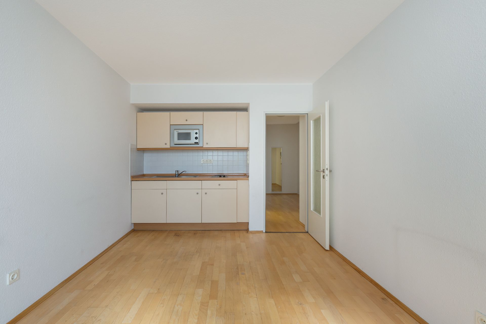 Helle Küche mit Einbauschränken, Spüle und Mikrowelle, angrenzend an einen Flur mit Holzboden.