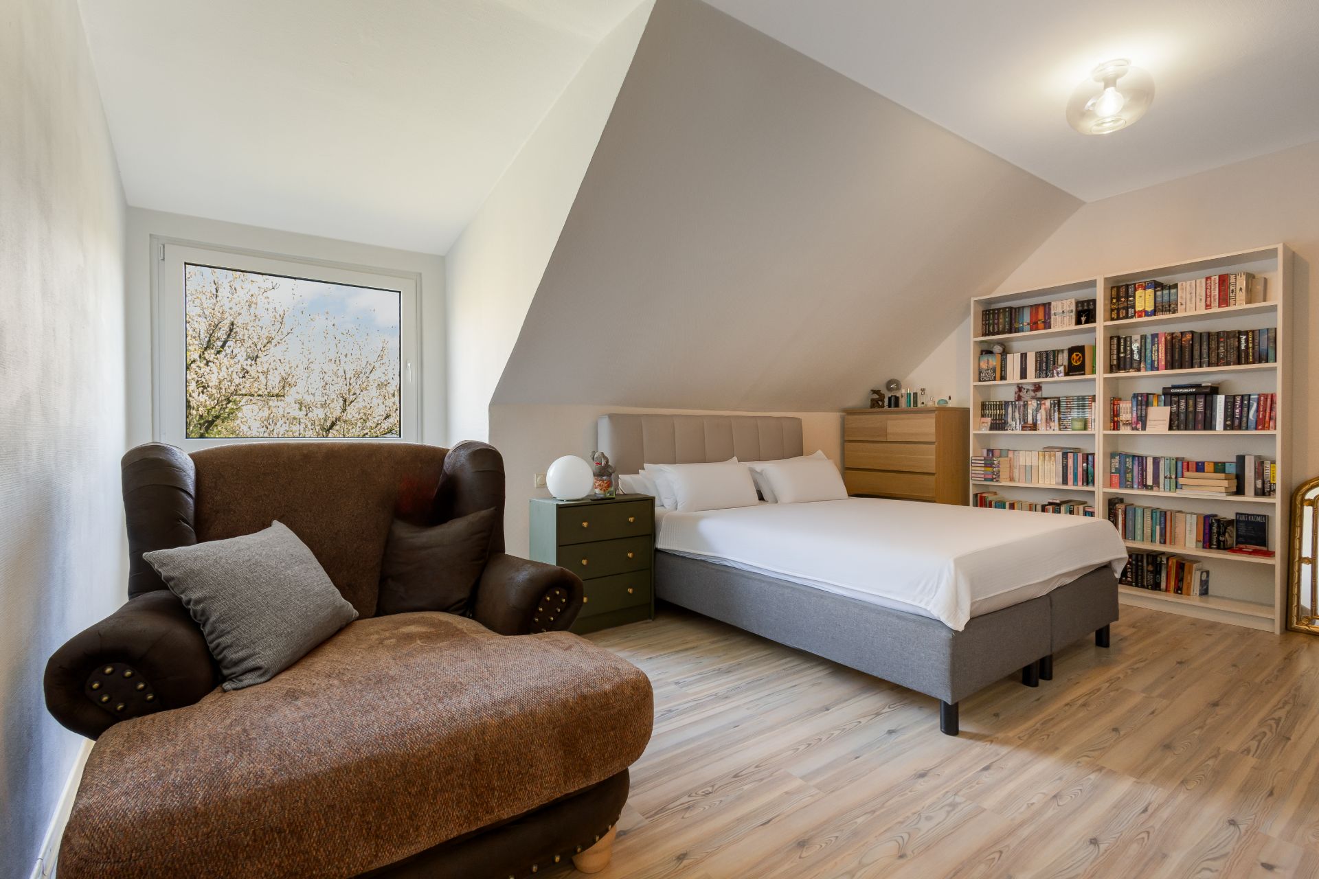 Helles Schlafzimmer mit Bett, Sessel, Bücherregal und Fenster mit Blick ins Freie.