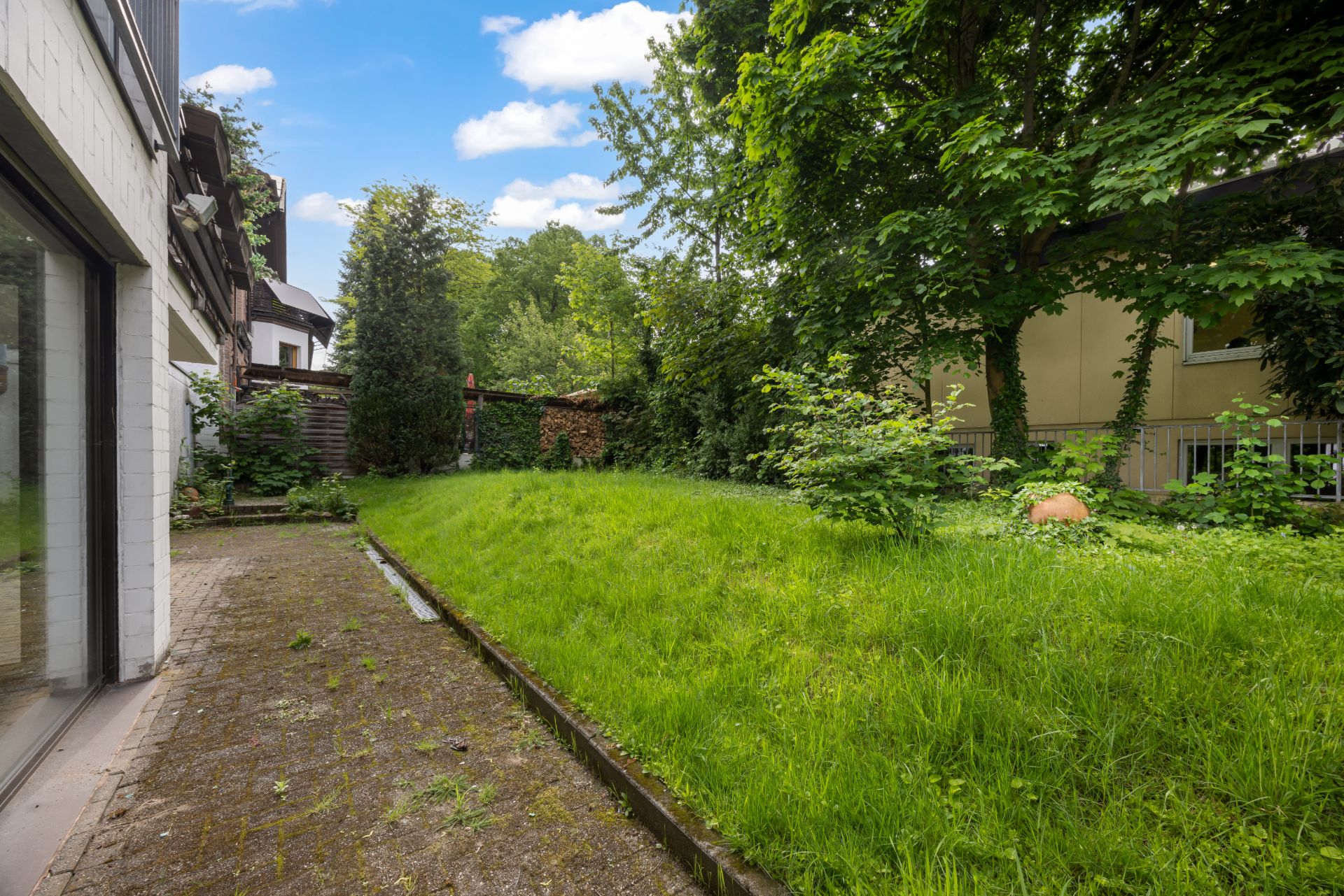 Garten mit Rasenfläche, Bäumen und gepflastertem Weg neben einem Wohnhaus.