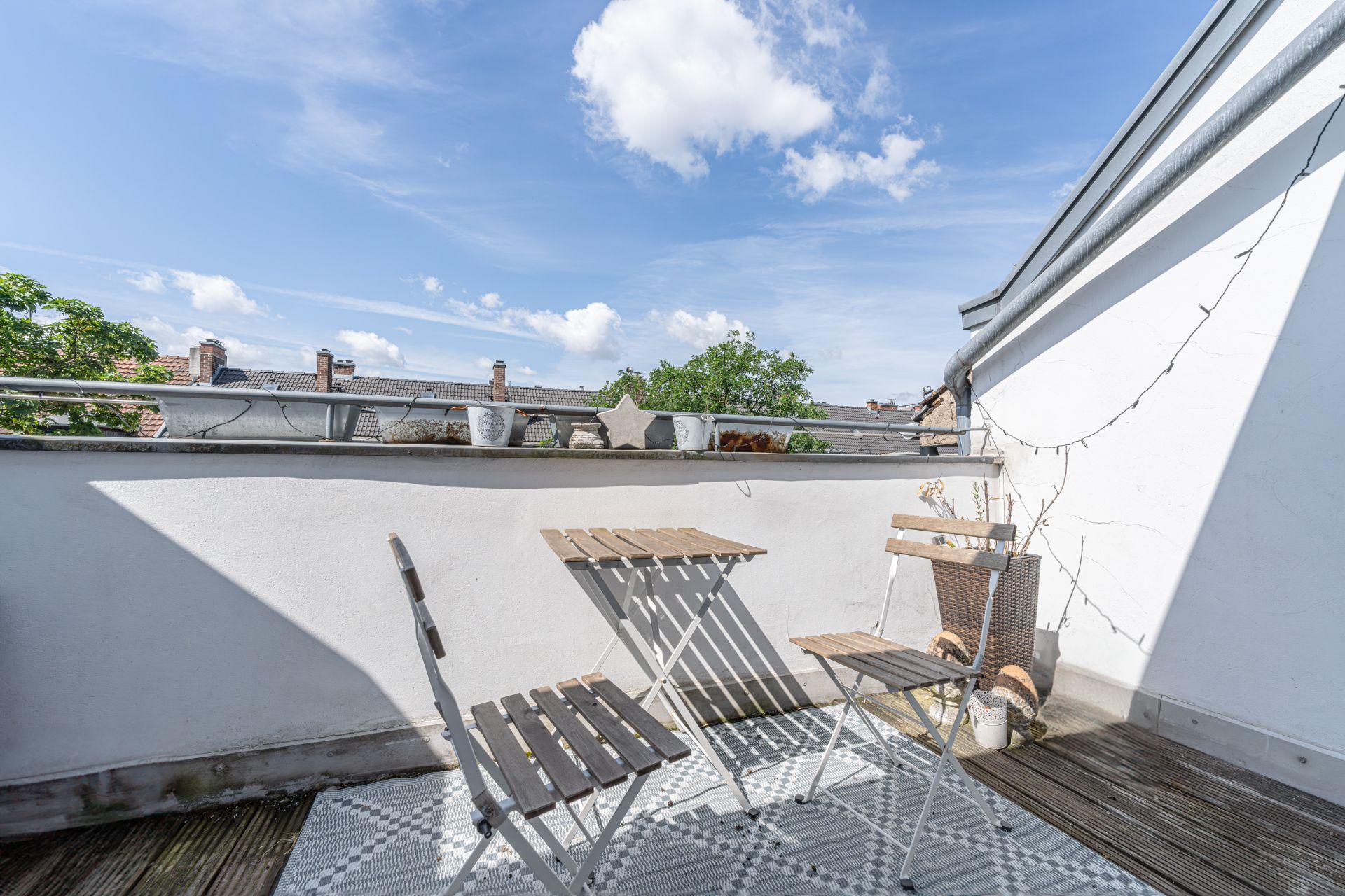 Kleine Terrasse mit zwei Stühlen, Tisch, Blumentöpfen und Lichterkette an einer weißen Wand.
