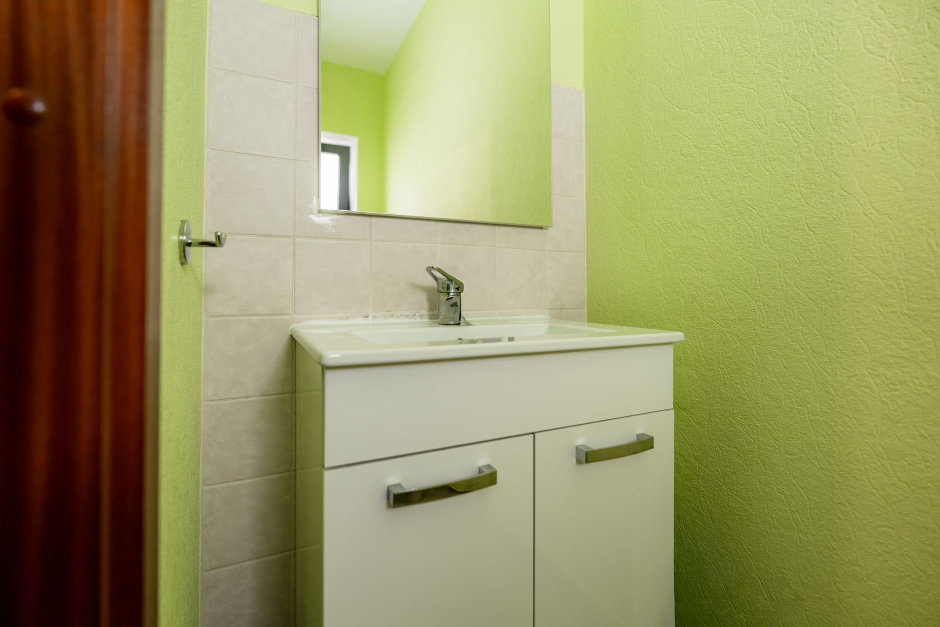 Waschbecken mit Unterschrank und Spiegel vor grüner Wand.