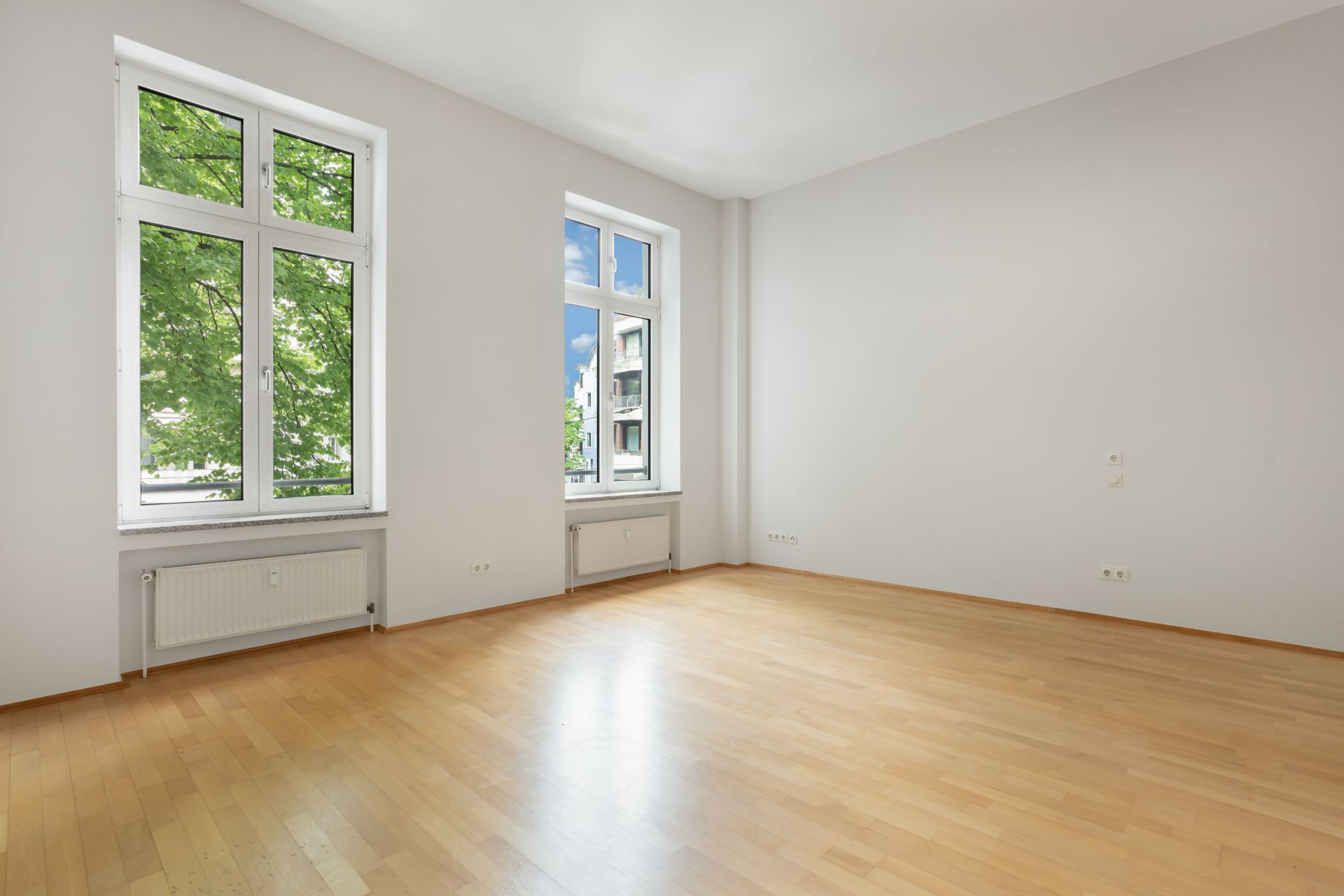 Helles, leeres Zimmer mit Parkettboden und zwei großen Fenstern.