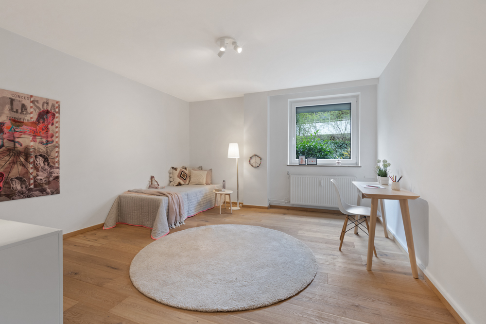 Helles Zimmer mit Einzelbett, Schreibtisch, rundem Teppich und Fenster mit Blick ins Grüne.