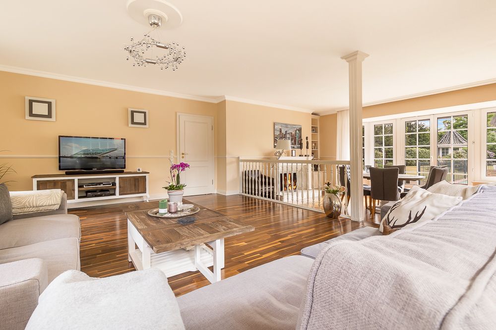 Helles Wohnzimmer mit Sofas, Holztisch, Fernseher und Essbereich mit großen Fenstern.