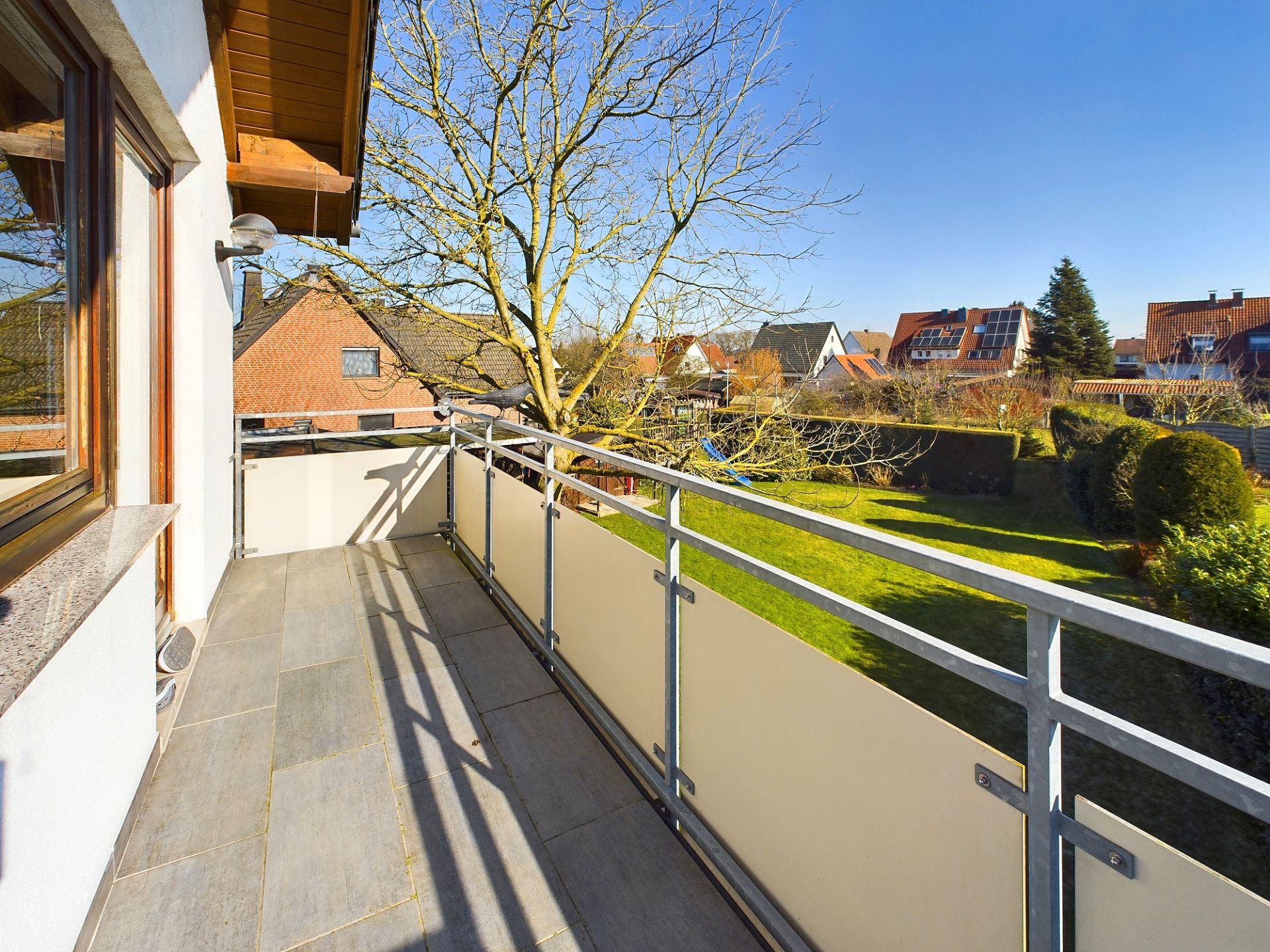 Balkon mit Metallgeländer und Blick auf Gärten und Wohnhäuser bei sonnigem Wetter.