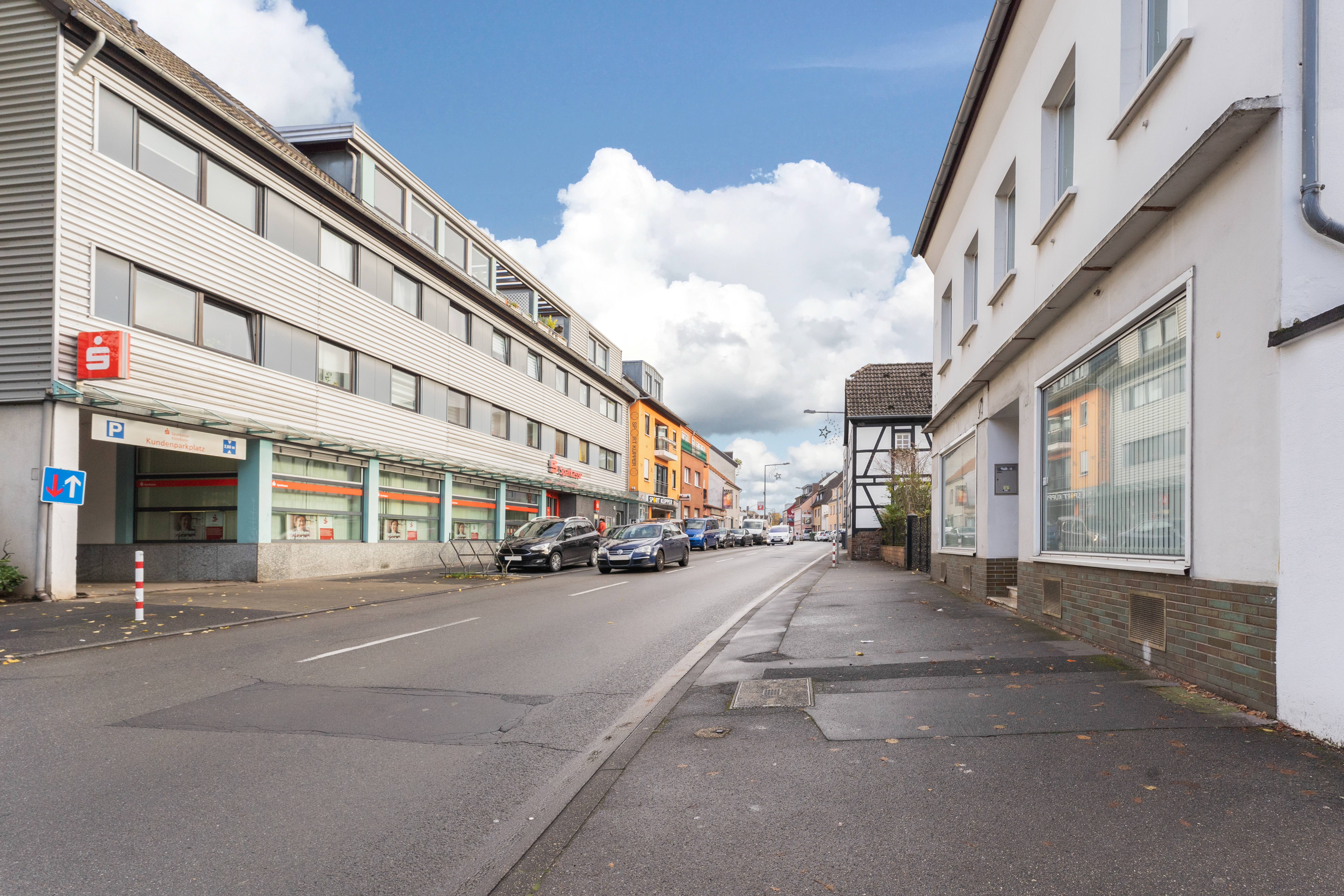 Blick auf eine ruhige Straße mit Wohn- und Geschäftsgebäuden, geparkten Autos und Gehweg.