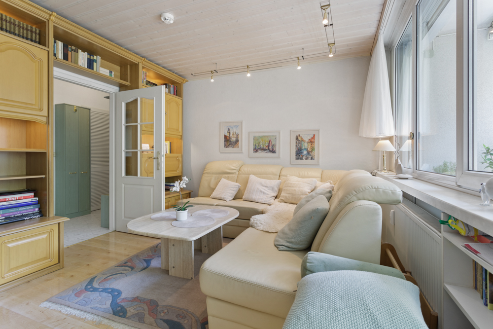 Helles Wohnzimmer mit Ecksofa, Couchtisch, Regalen und großen Fenstern.