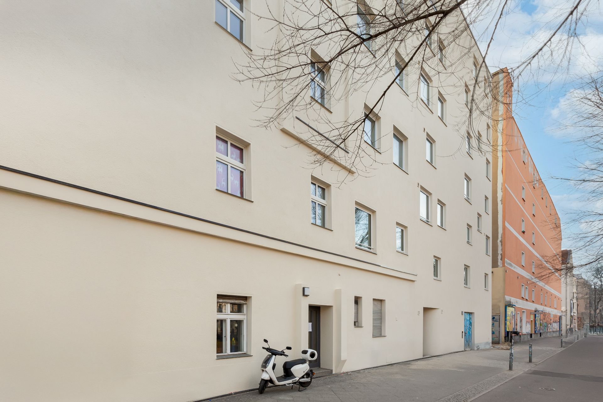 Helles Wohnhaus mit mehreren Fenstern, ein Roller steht vor dem Eingang auf dem Bürgersteig.