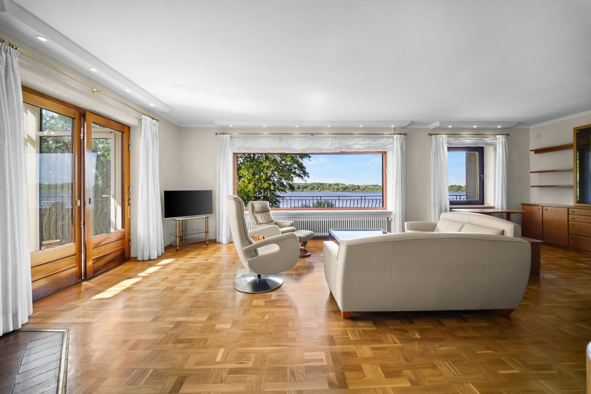Helles Wohnzimmer mit weißen Möbeln, Parkettboden und großen Fenstern mit Seeblick.