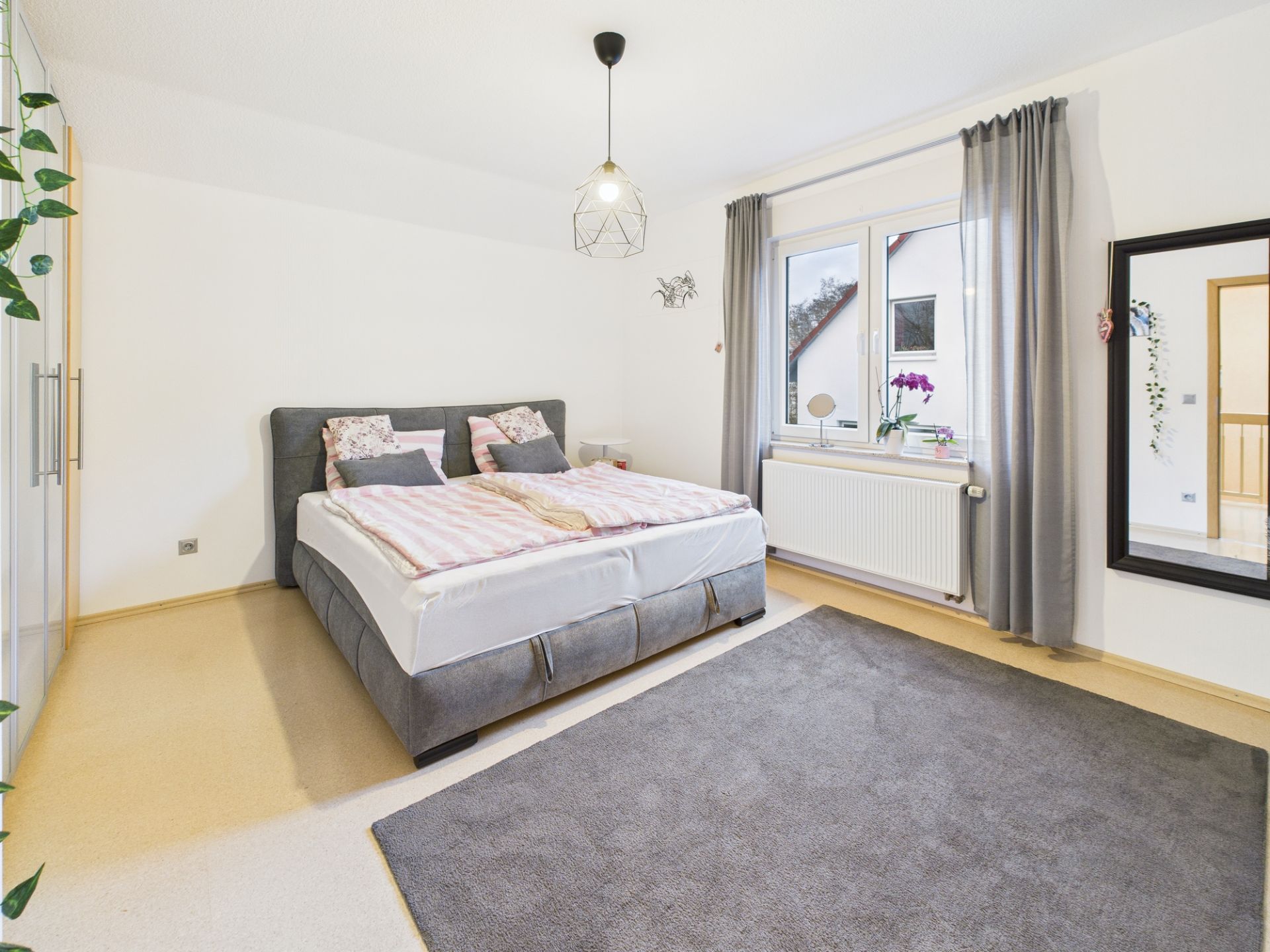 Helles Schlafzimmer mit Doppelbett, großem Fenster, grauem Teppich und Spiegel an der Wand.