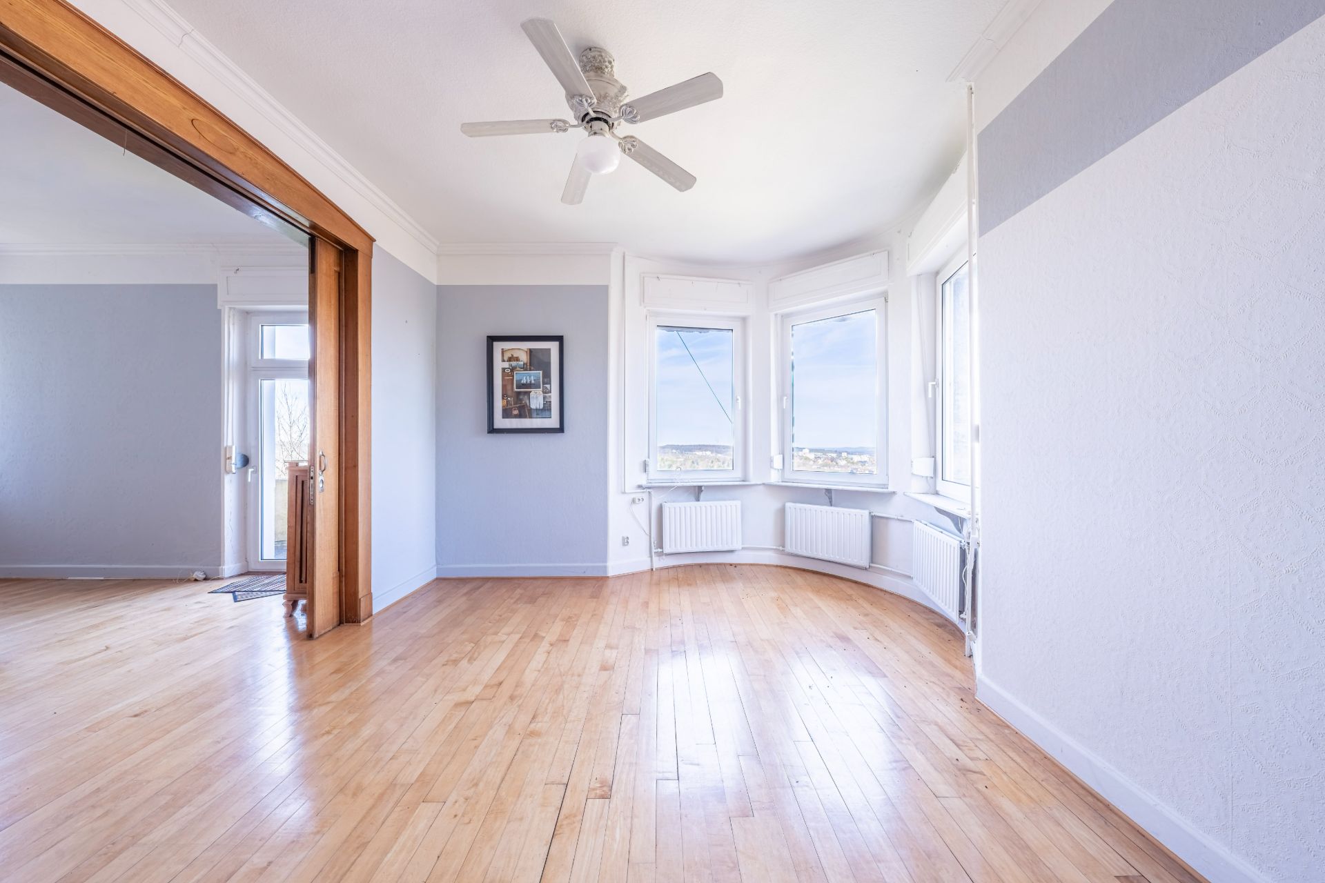Heller Raum mit Holzboden, Deckenventilator, großen Fenstern und einem Wandbild.