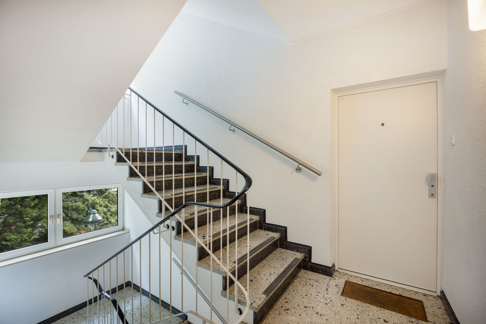 Treppenhaus mit Handlauf, Fenster und weißer Wohnungstür auf der rechten Seite.