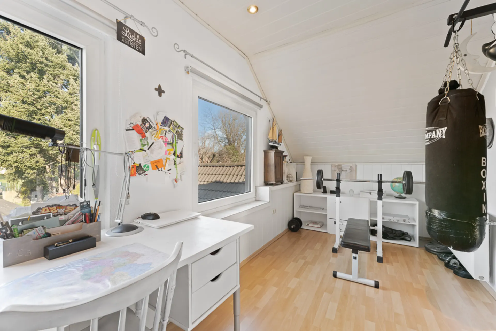 Helles Zimmer mit Schreibtisch, Hantelbank, Boxsack und Regal unter Dachschräge.